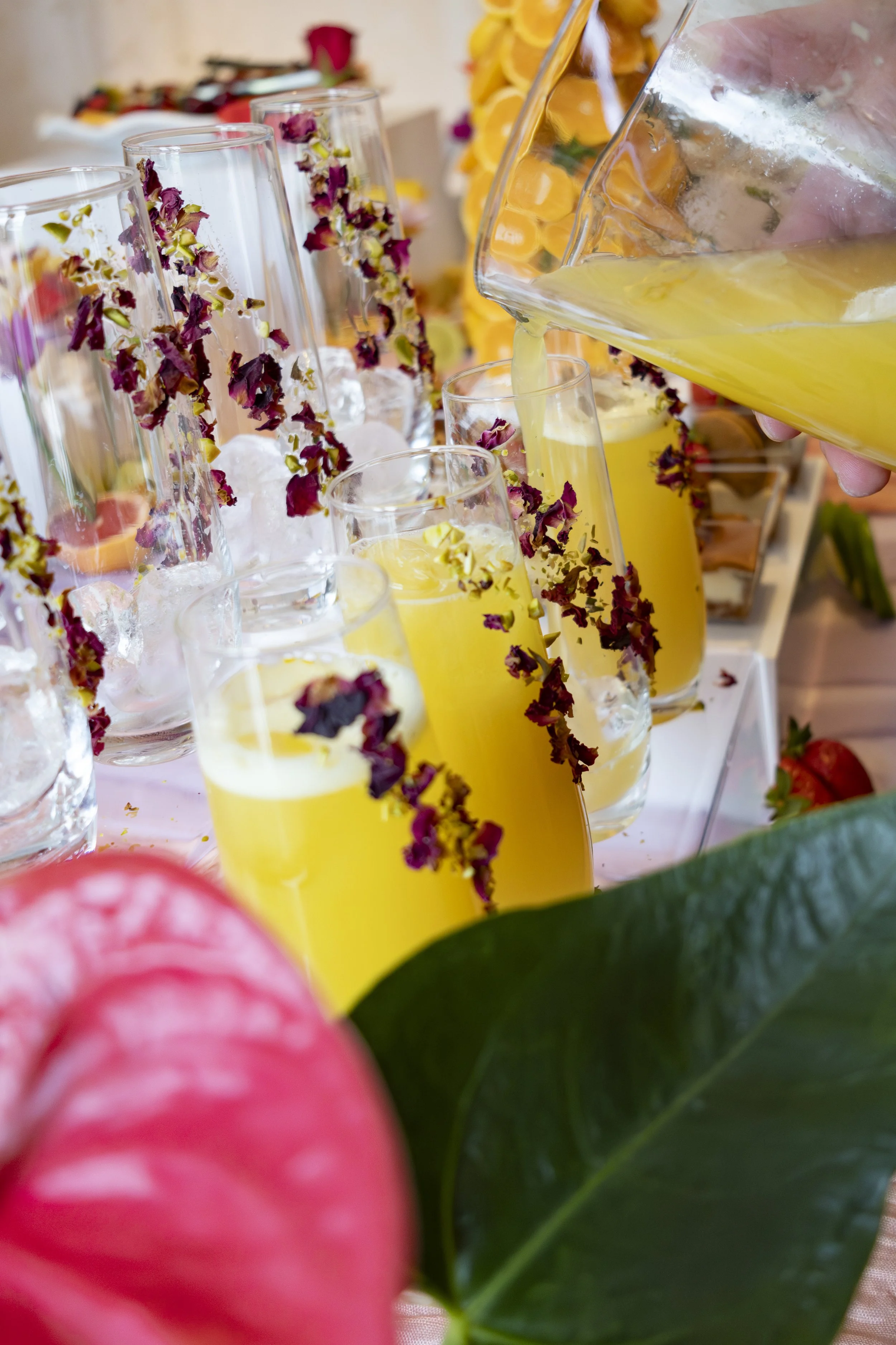 Close-up view of a beverage station with yellow drinks in tall glasses, garnished with dried flower petals, and a pitcher pouring more of the yellow drink. In the background, there are decorated glasses with ice and flower petals, as well as decorati