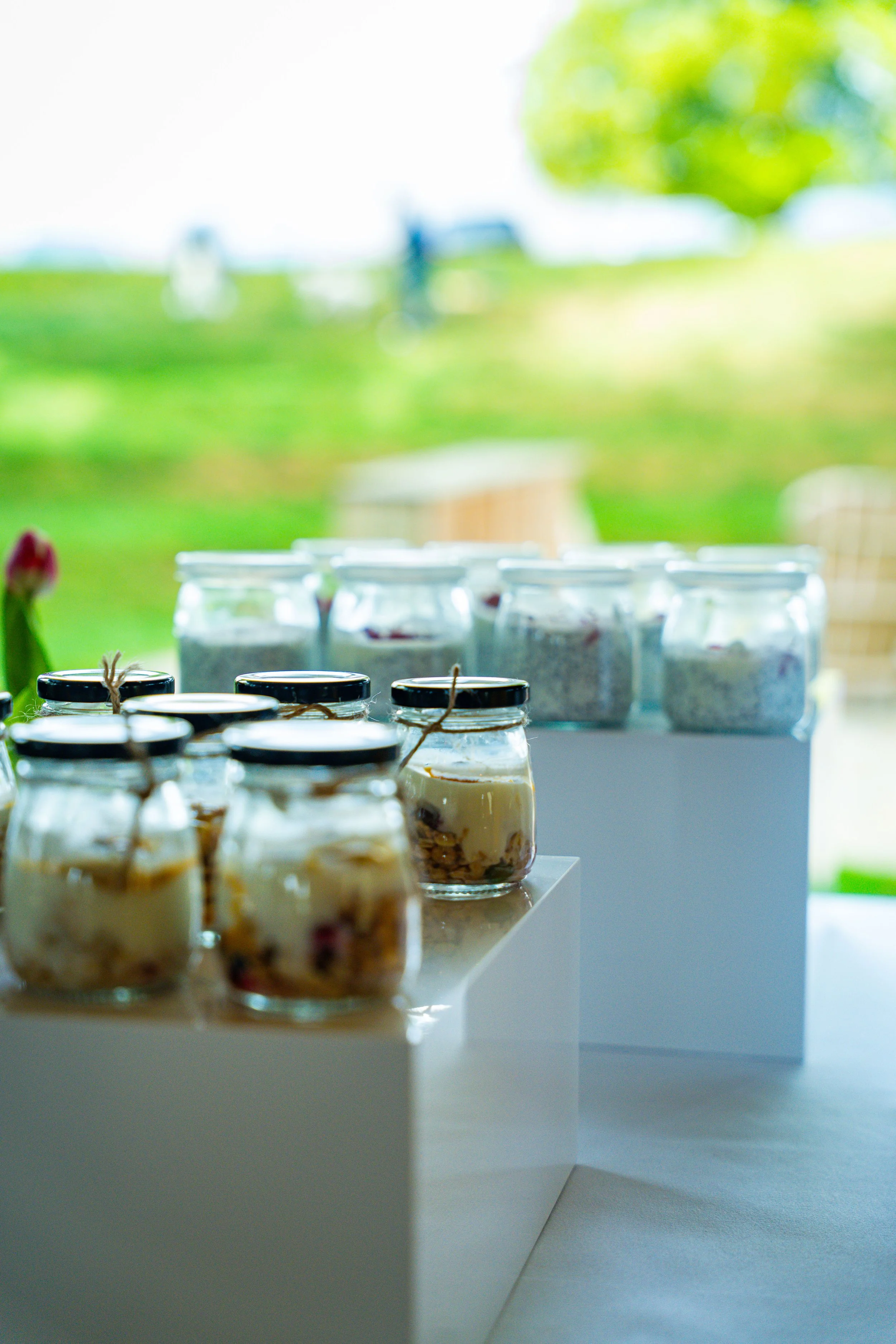 Display of glass jars with desserts, possibly parfait or yogurt, on a white stand in a bright, airy setting with green outdoor scenery visible through a large window.