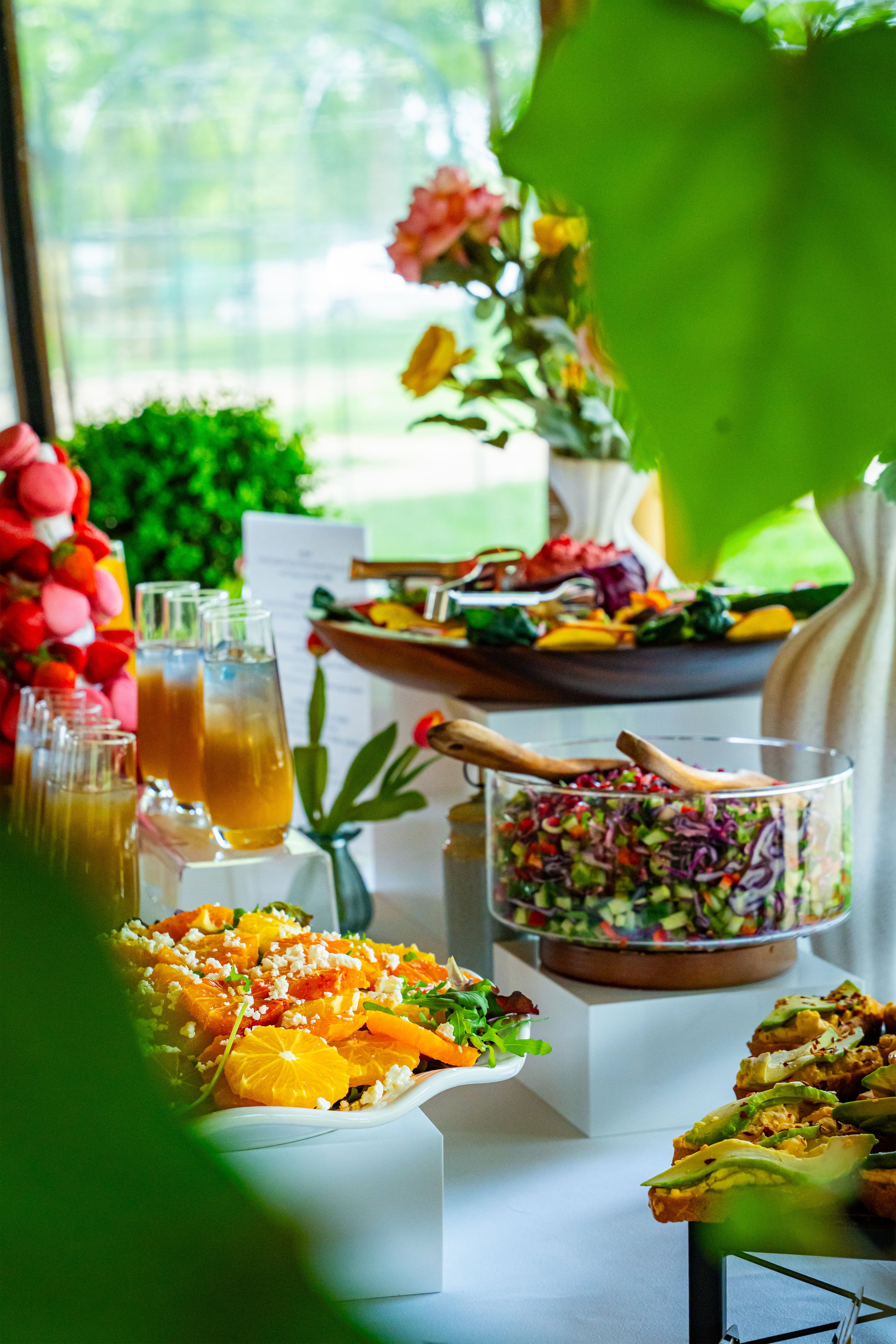 Table with a variety of colorful salads and desserts, decorated with flowers, and set in a bright room with large windows and greenery outside.