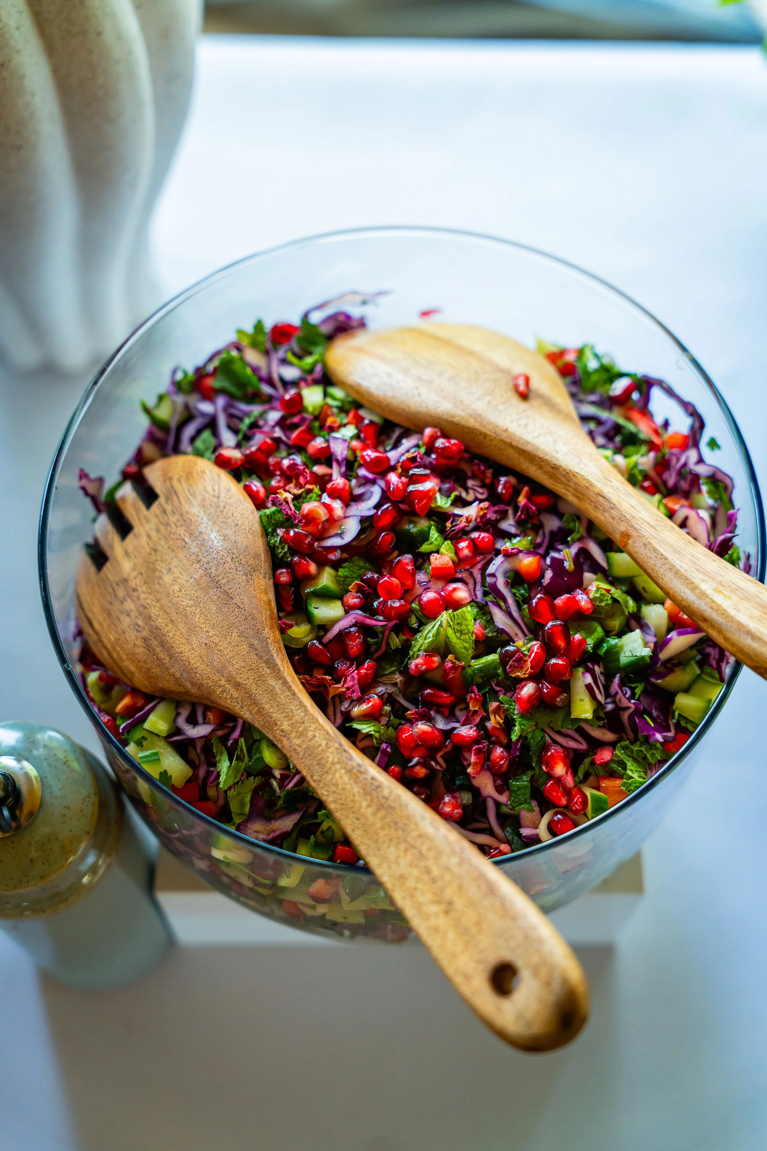 A glass bowl of chopped vegetables and pomegranate seeds with two wooden salad servers inside, placed on a white surface.