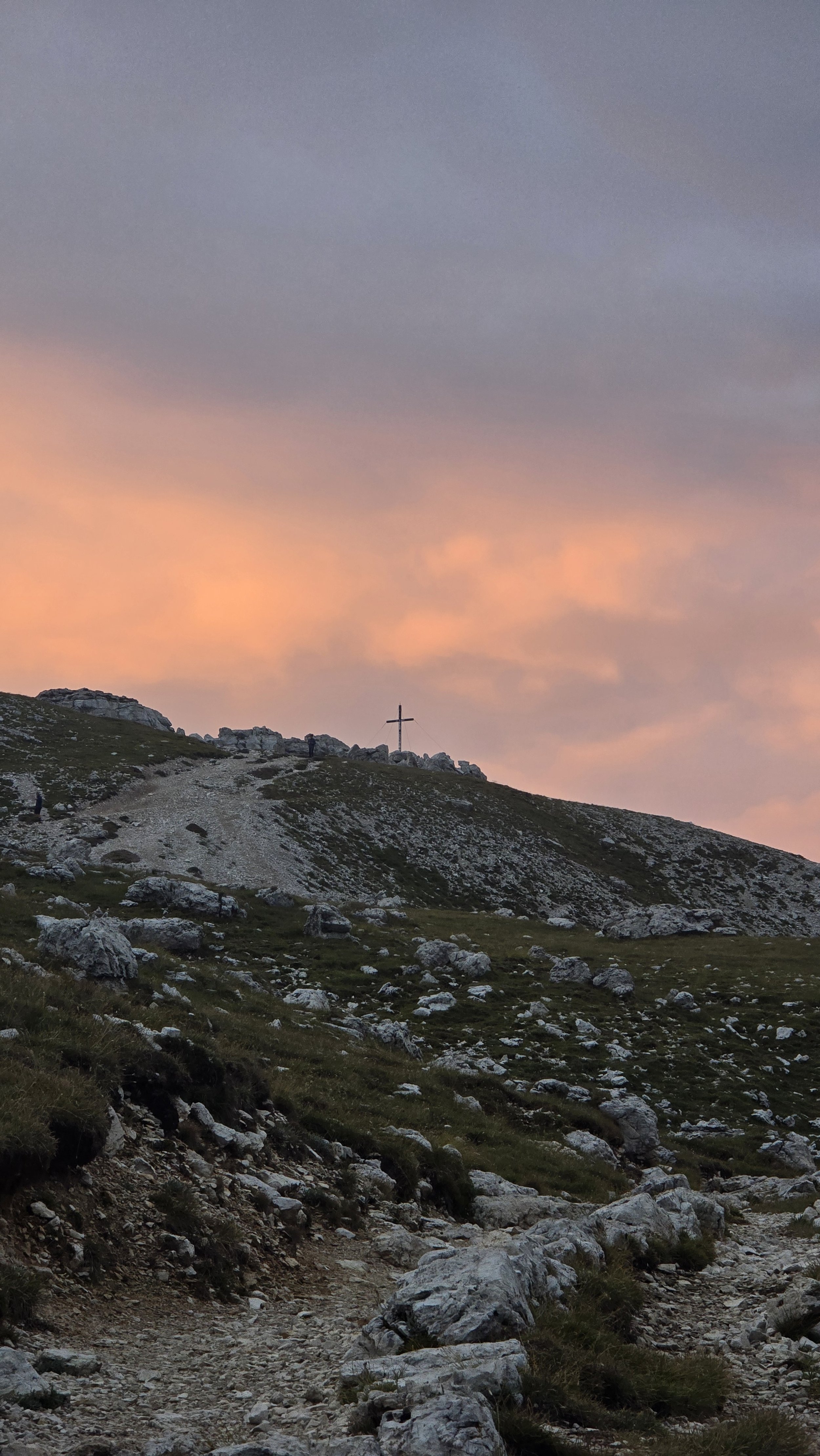 A mountain trail leading up a rugged rocky hillside during sunset, with a cross at the top of the hill and a few people walking along the path.