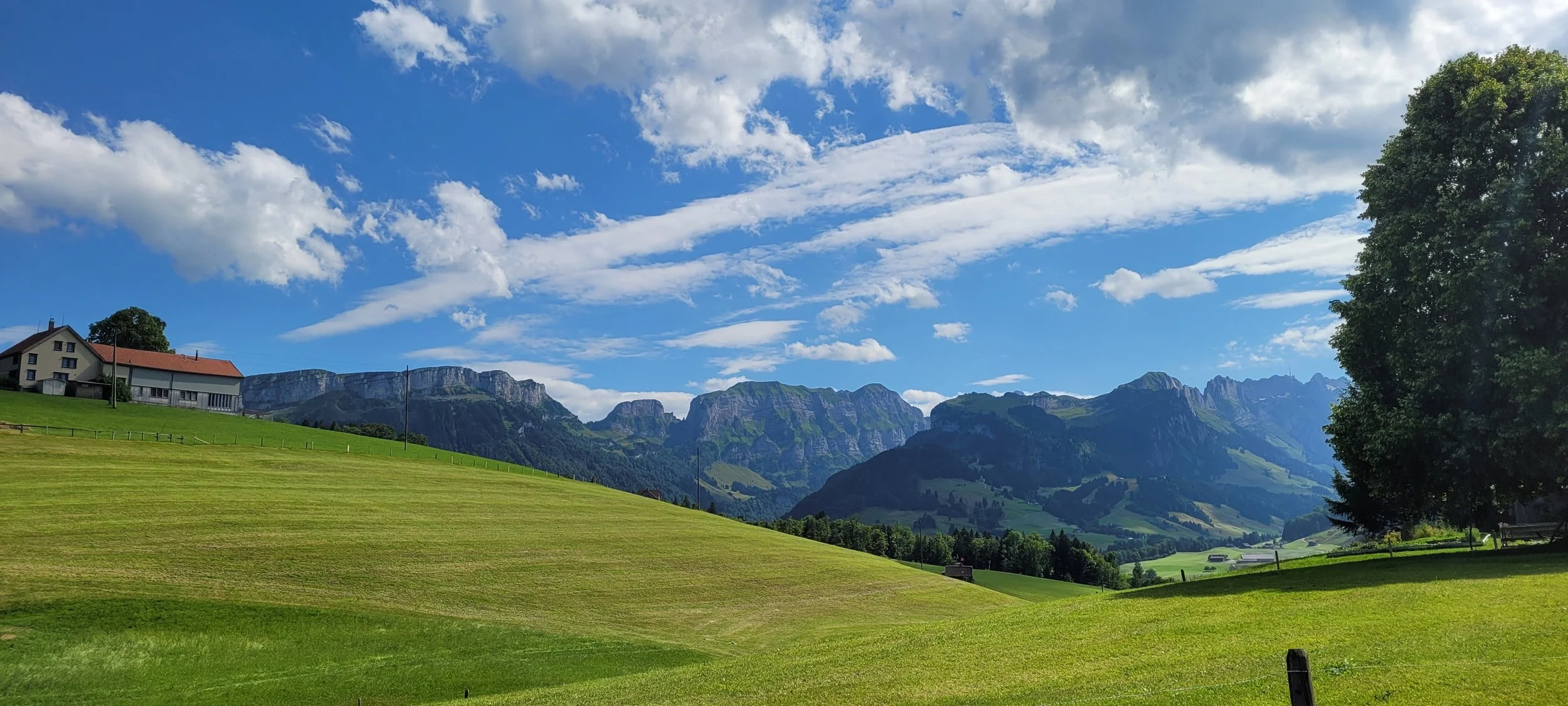 A scenic landscape with green rolling fields, a few farm buildings, a large tree on the right, distant mountains, and a partly cloudy blue sky.
