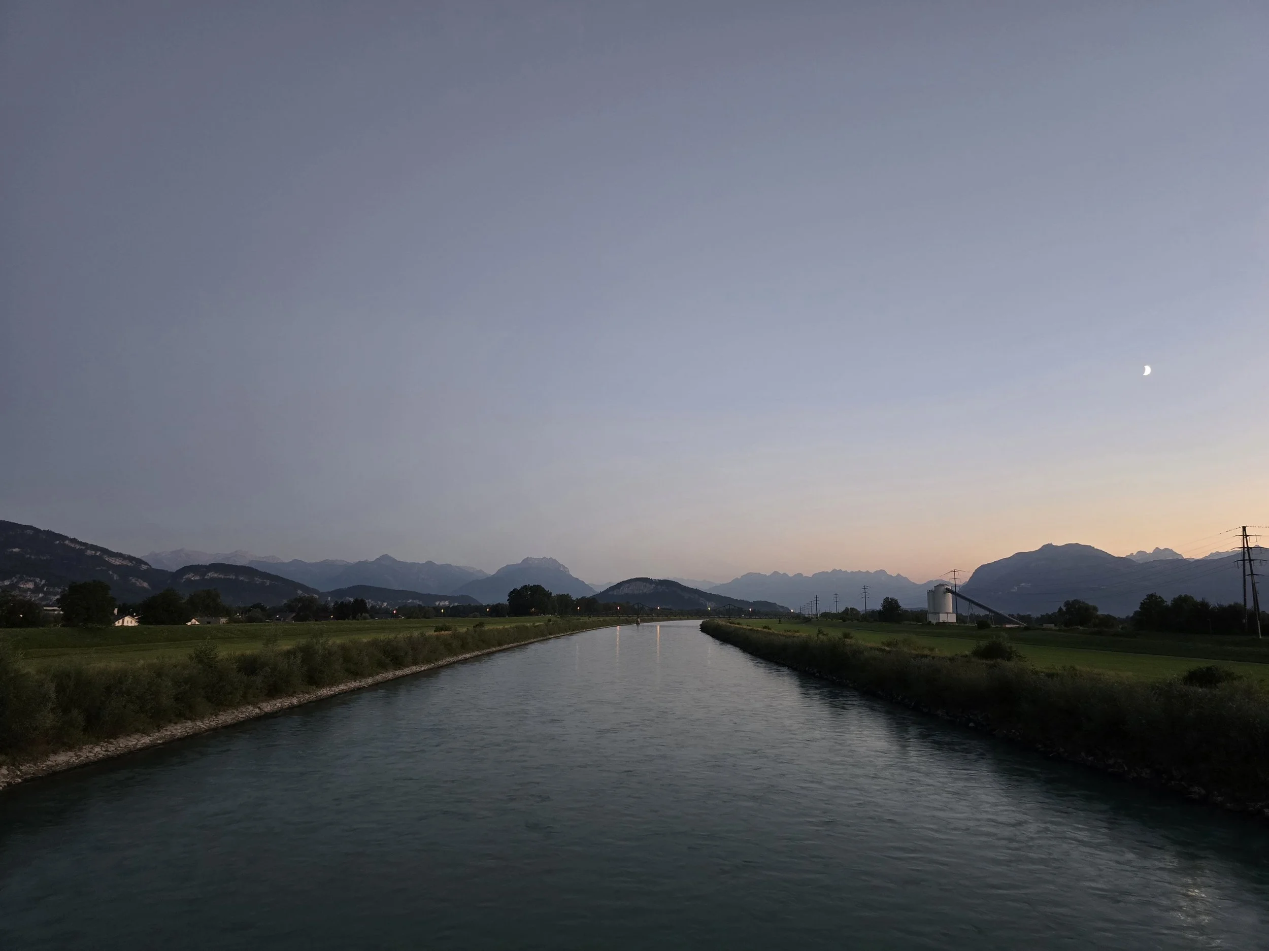 A calm river reflecting the evening sky with a crescent moon, surrounded by green fields, with mountains in the background and a few industrial structures, including power lines and a silo.