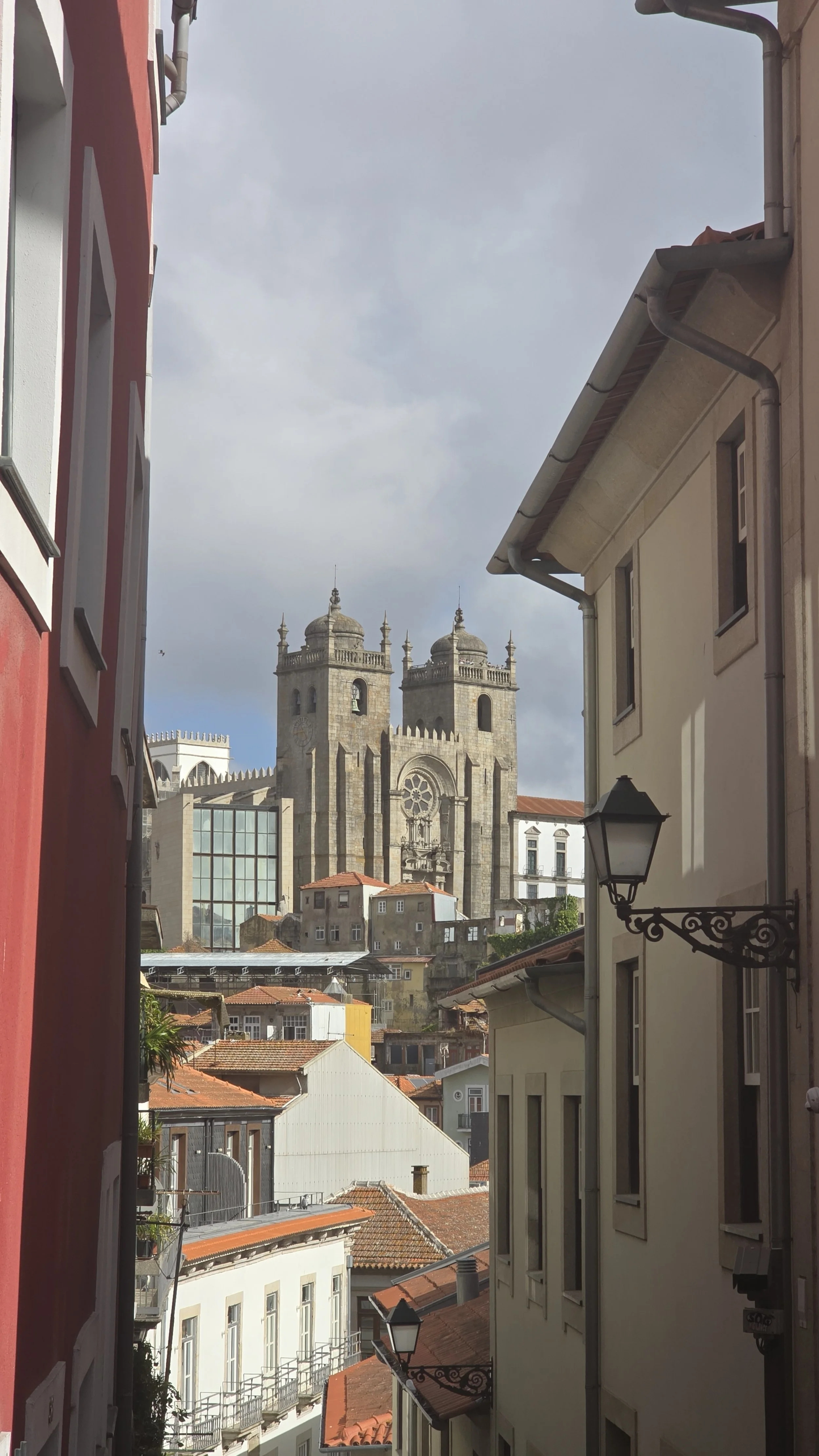 View of a historic stone church with twin towers seen from a narrow street lined with colorful buildings with red-tiled roofs.