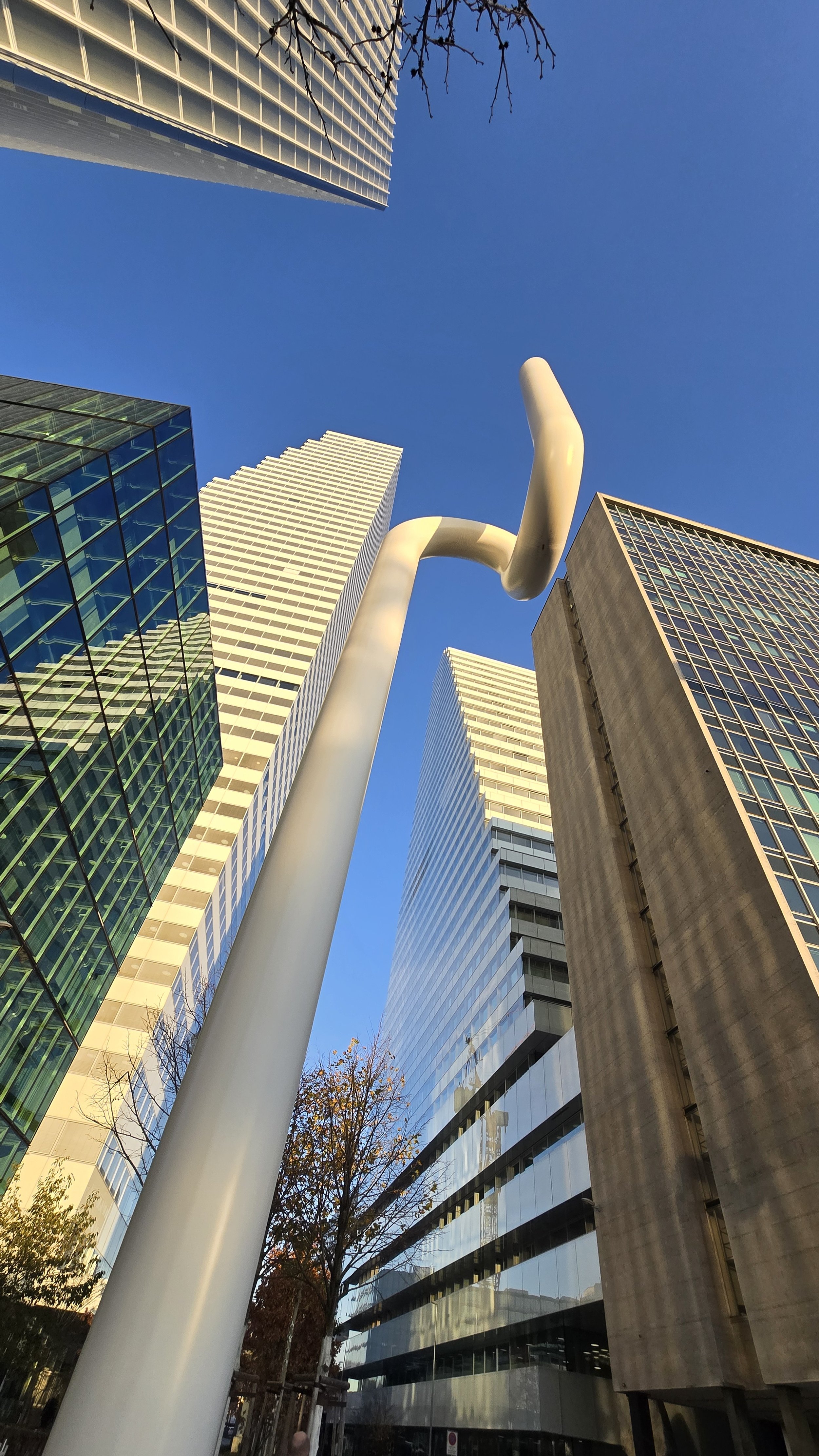 Low-angle view of tall modern skyscrapers with glass facades and a unique white streetlamp against a clear blue sky, with trees at the bottom of the scene.