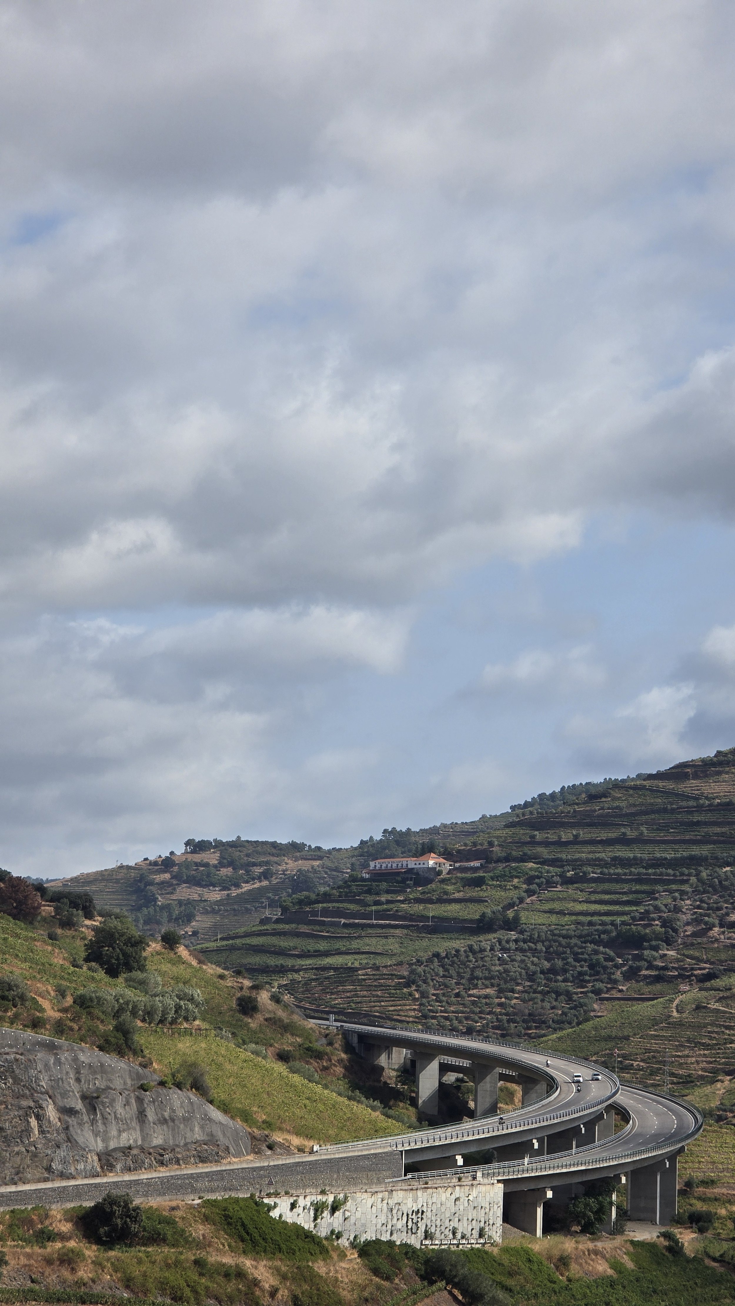 A winding highway on hilly terrain with terraced farmland, trees, and a building on one of the hills, under a partly cloudy sky.