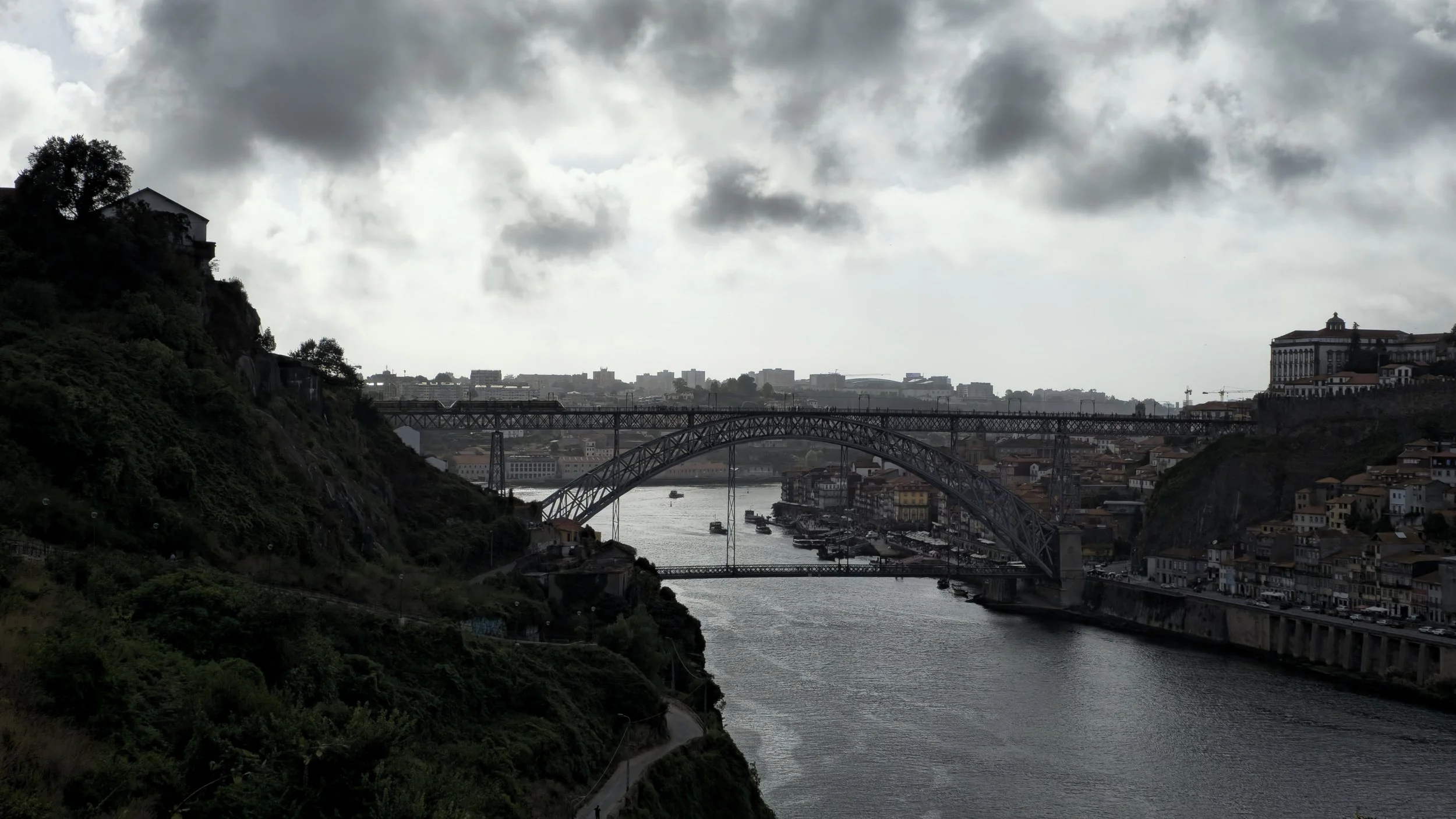 A river flowing through a city with multiple bridges, surrounded by hills with buildings on both sides, under a cloudy sky.