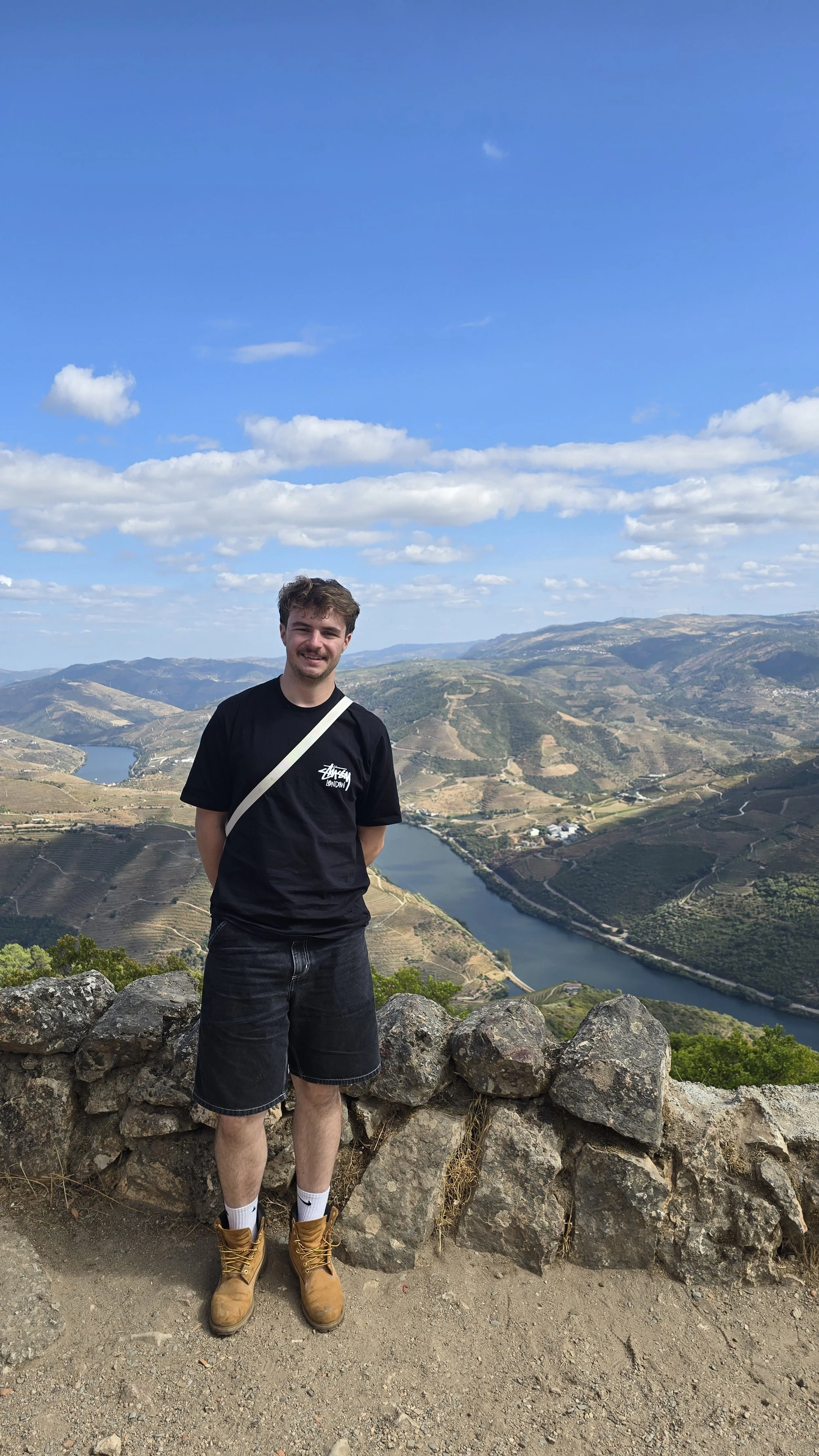 A young man standing on a rocky ledge with mountains, a river, and a blue sky with scattered clouds in the background.