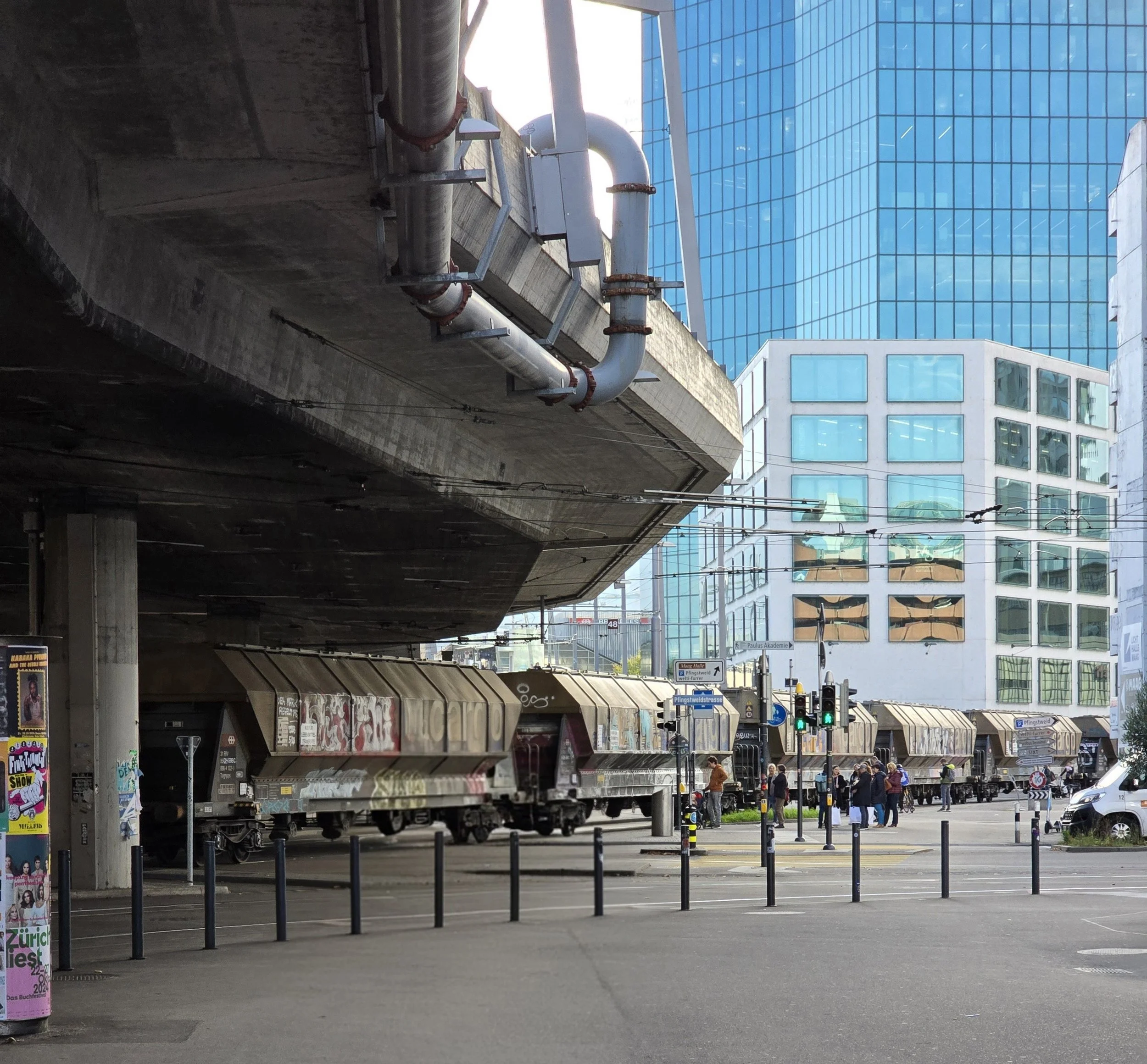 City scene with train cars parked under an overpass, multiple pedestrians crossing at the intersection, modern glass buildings, traffic lights, and overhead tram wiring.