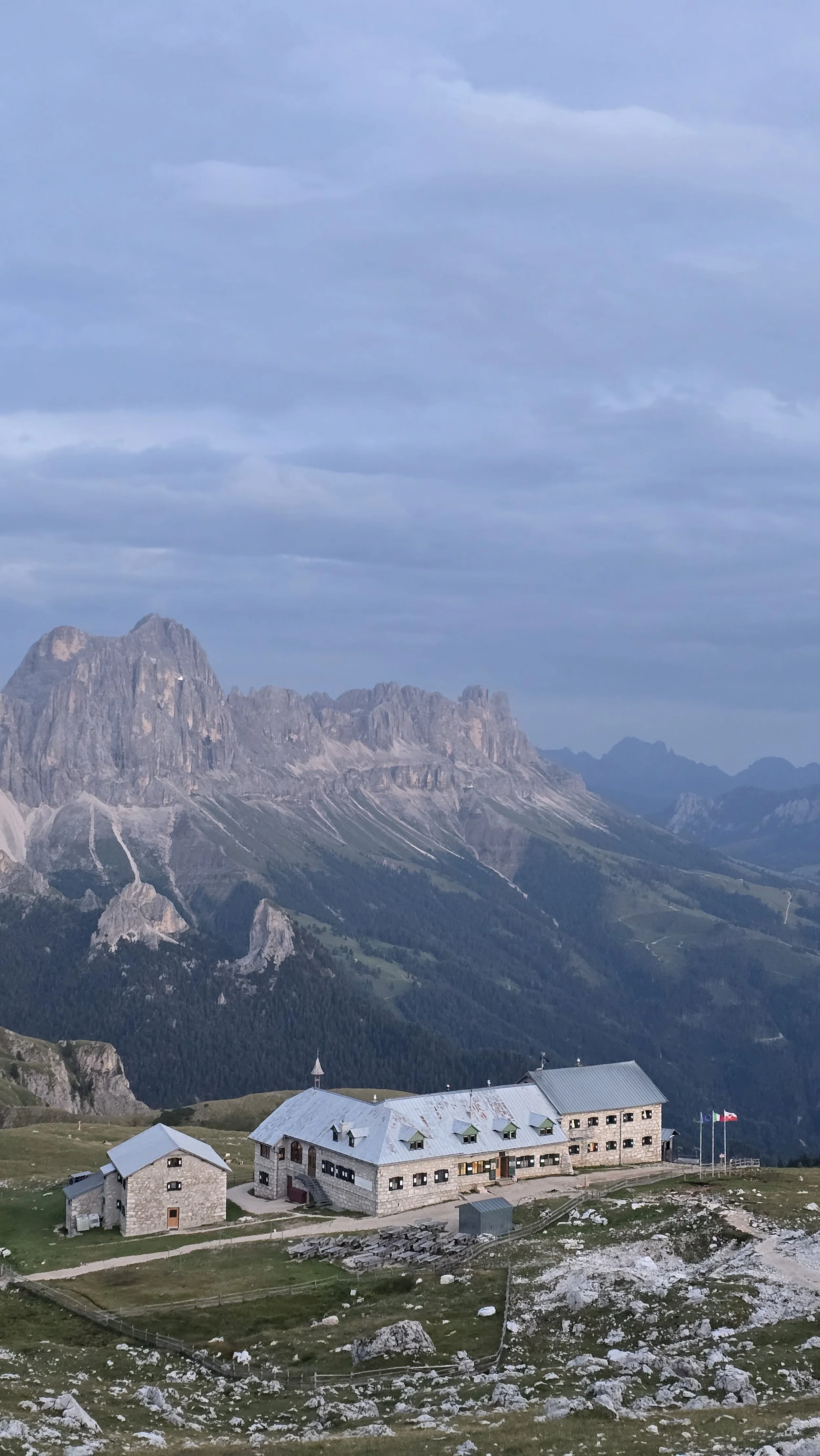 Mountain landscape with rocky peaks in the background, a rustic stone building with a metal roof in the foreground, and a dirt path leading to the building, under a cloudy sky.