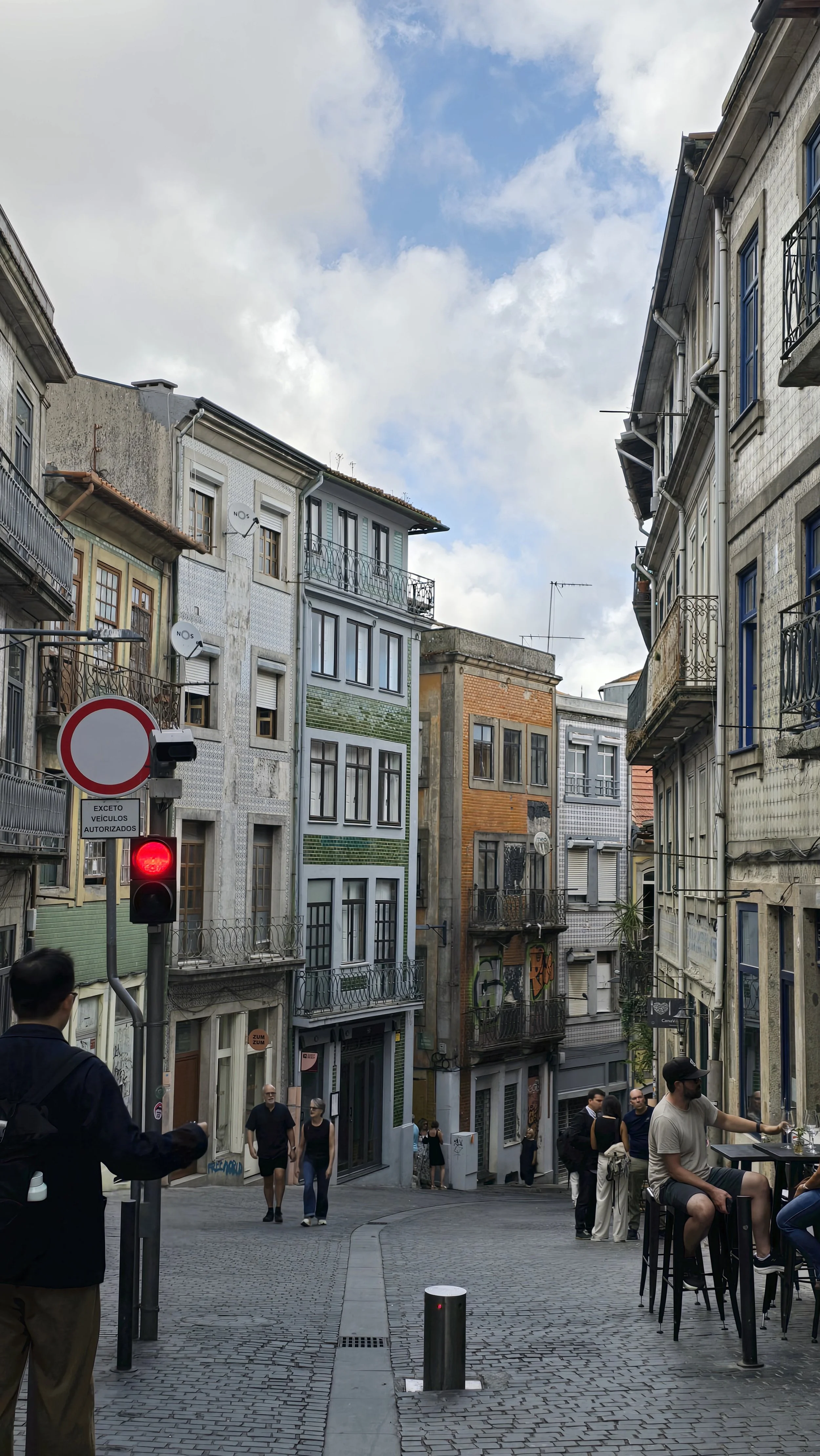 A cobblestone street in an urban area with tall, colorful apartment buildings on both sides. Several pedestrians are walking or sitting, including a man with a backpack and a woman with sunglasses. There are outdoor seating areas at a cafe on the right, and a traffic light showing red with a no-entry sign and a sign in Portuguese indicating it's for authorized vehicles only.