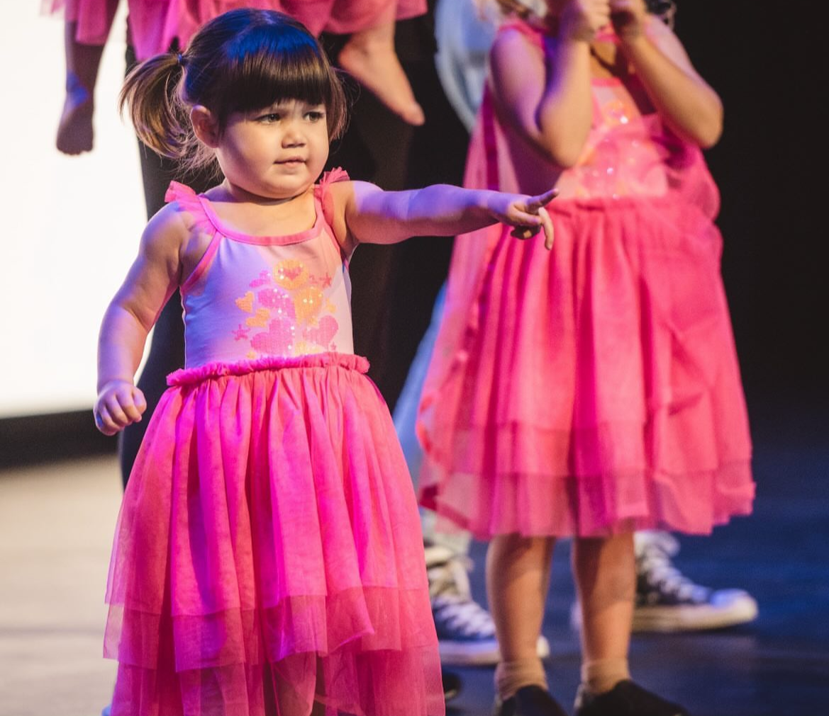 A young girl in a pink dance costume points forward during a performance on stage.