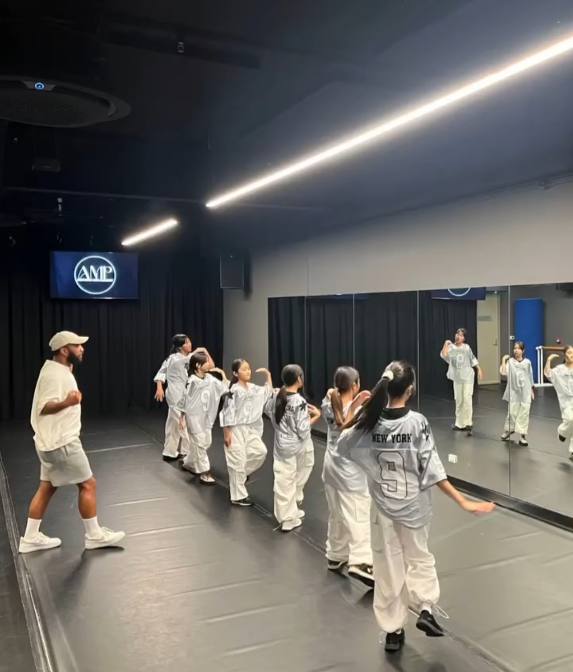 Children are in a dance studio practicing moves in front of a mirrored wall, led by an instructor. The children are wearing sports uniforms with 'New York' on the back.