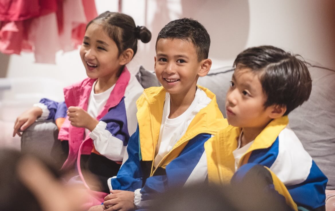 Three children sitting on a sofa, smiling and looking at something off to the left.