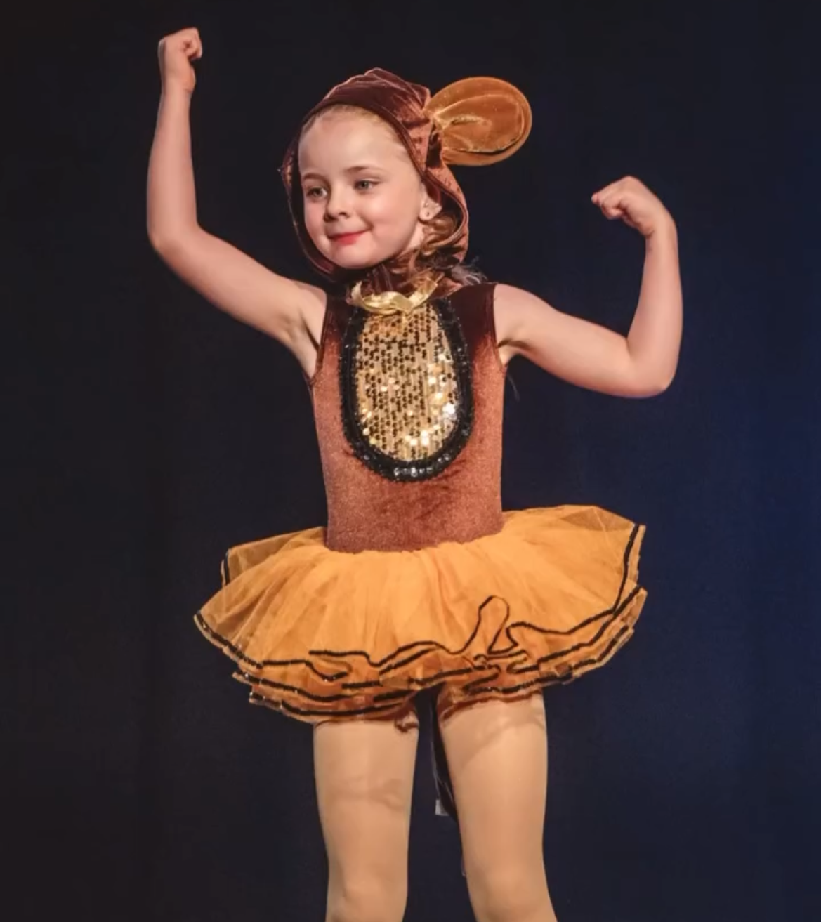 A girl dressed in a shiny orange ballet costume with a tutu, flexing her arms, and wearing a headpiece resembling a peanut.