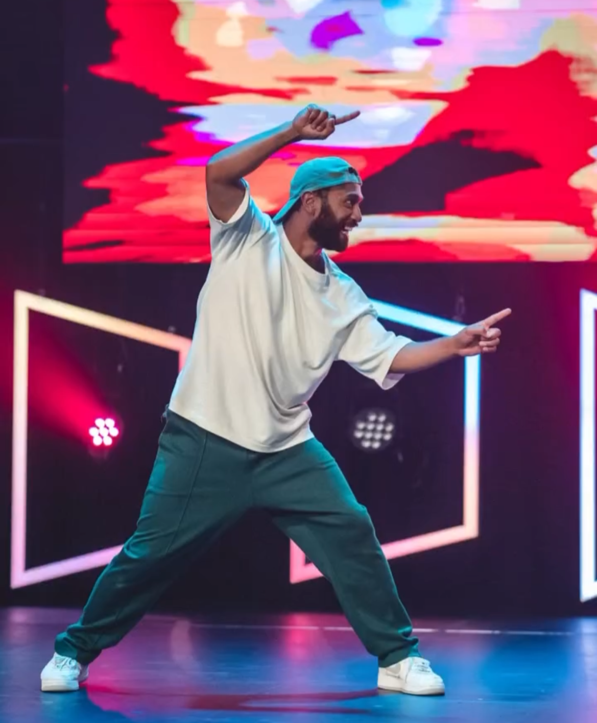 A man with a beard, wearing a light-colored oversized t-shirt, dark pants, white sneakers, and a backward baseball cap, dancing on a stage with colorful neon lights and abstract digital backgrounds.