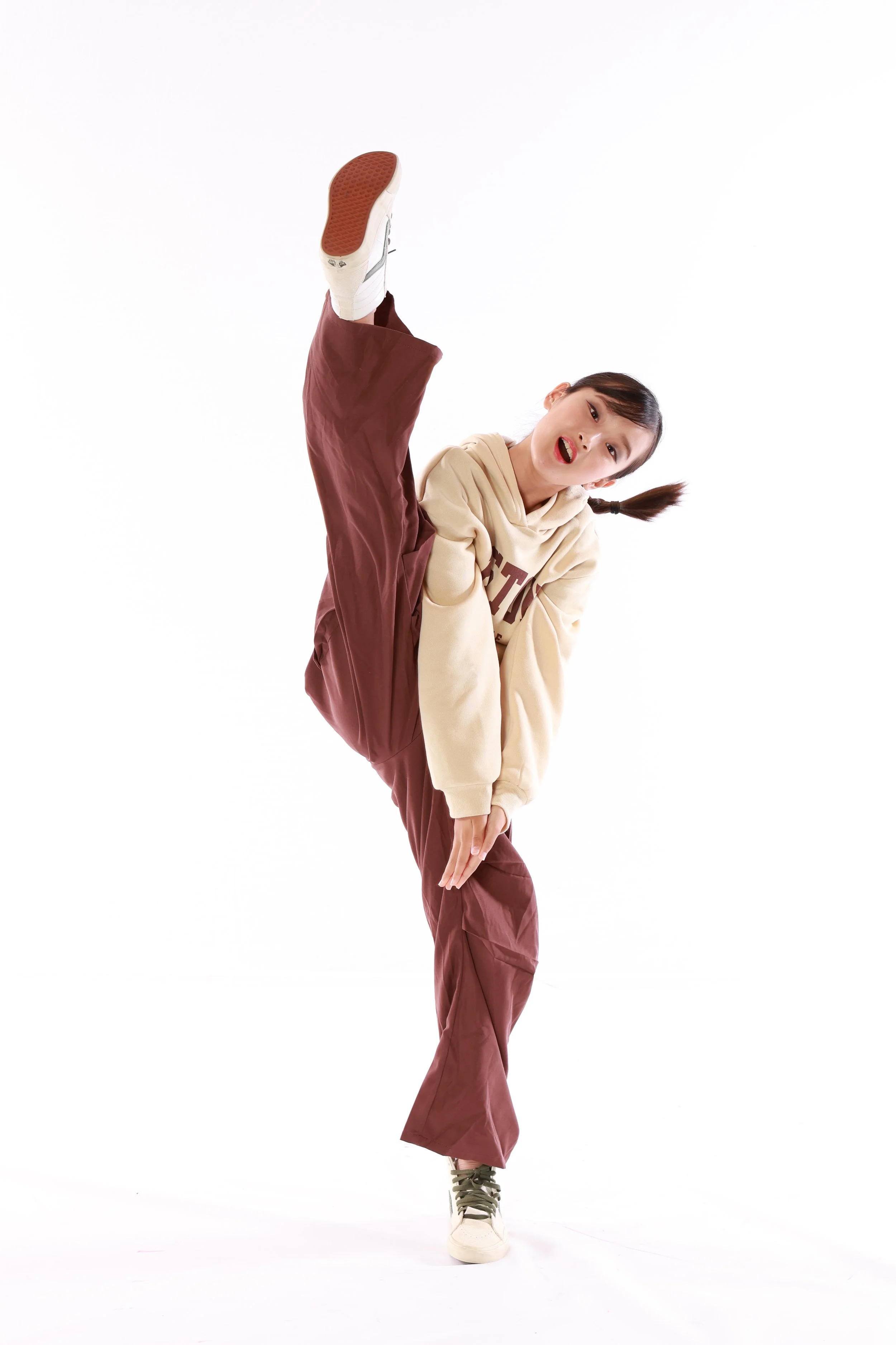 A young woman performing a high kick, wearing beige hoodie, maroon loose pants, and white sneakers, against a white background.