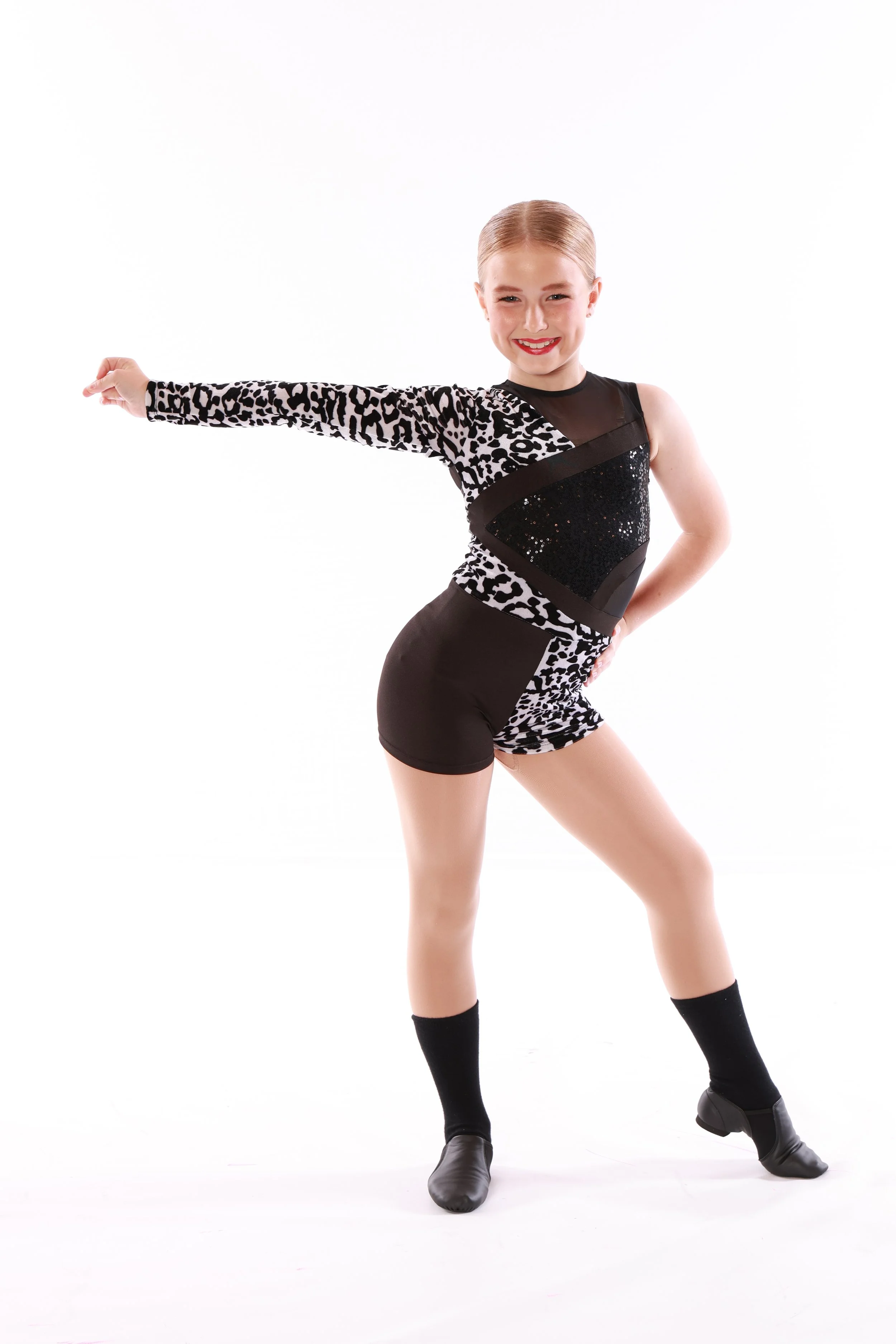 Young girl in a black and leopard print dance costume, performing a dance pose with one arm extended and smiling.