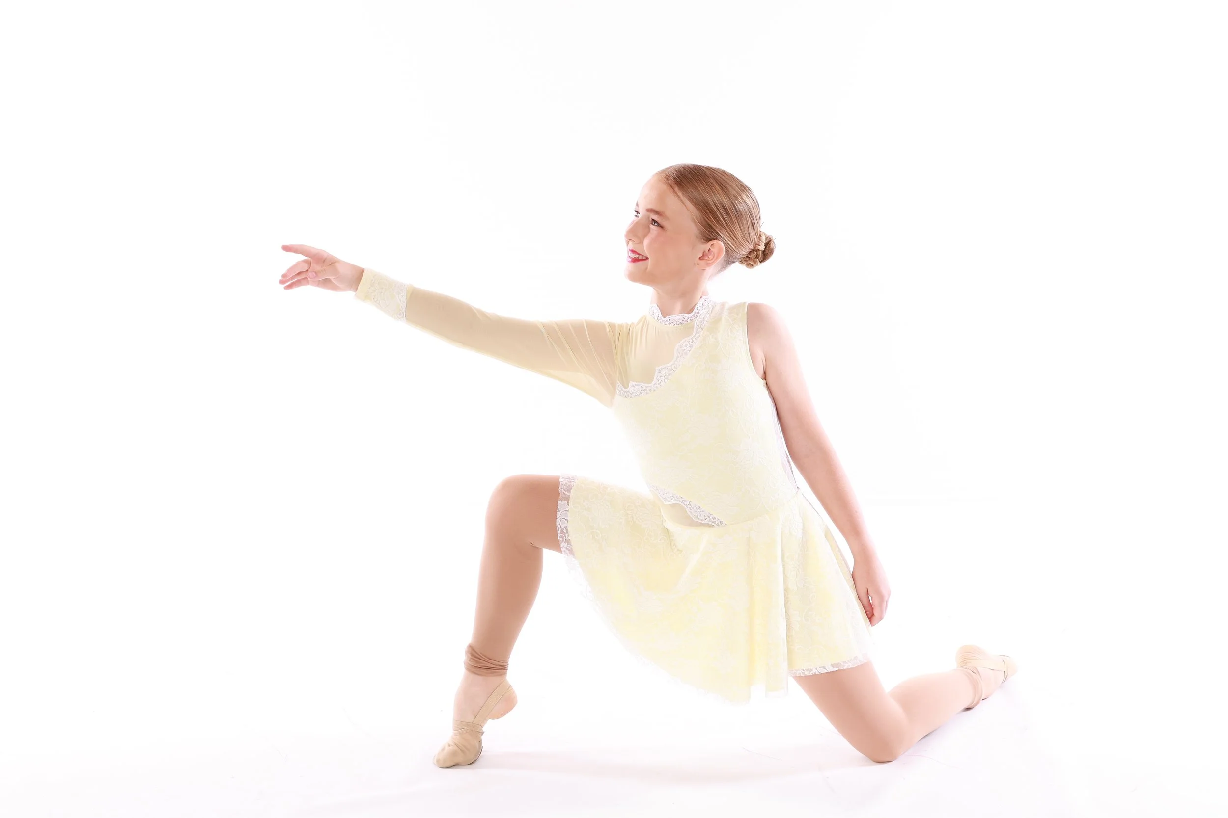 A young woman in a yellow ballet dress and tan pointe shoes is performing a ballet pose on a white background, extending one arm forward and kneeling on one knee.