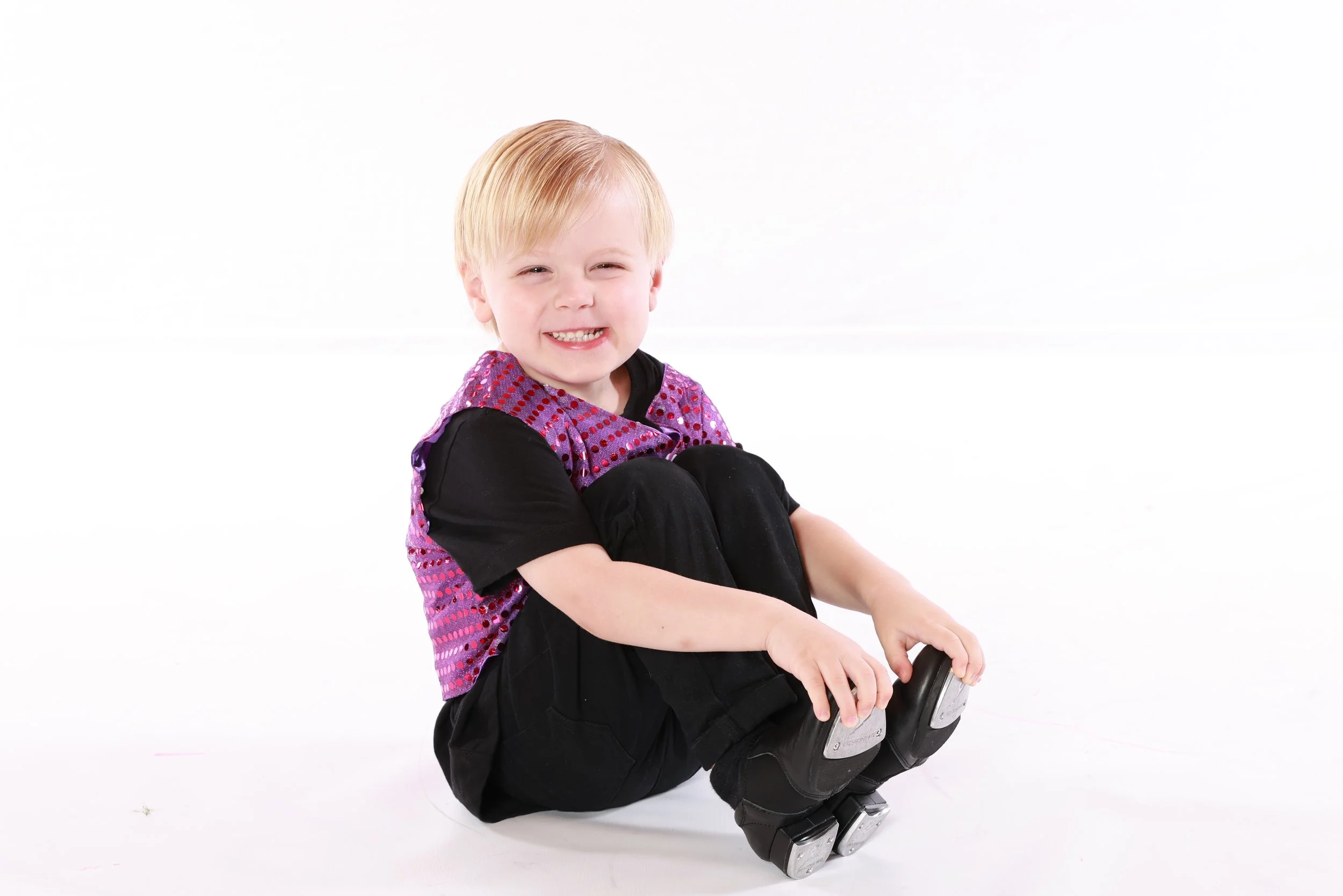 A young boy with blonde hair sitting on the floor, smiling, wearing black pants, a black t-shirt, and a purple and pink vest, holding his rollerblades.