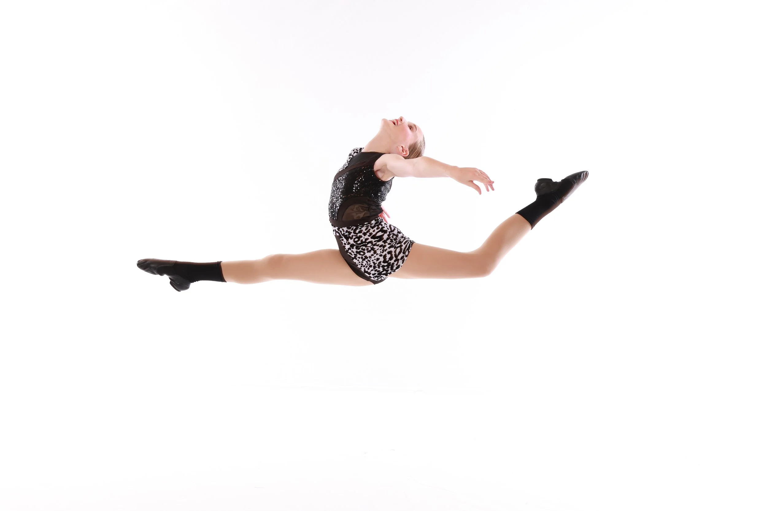 A female dancer performing a split leap with her arms extended backward, wearing a black top with sequins, leopard-print shorts, and black shoes, against a white background.