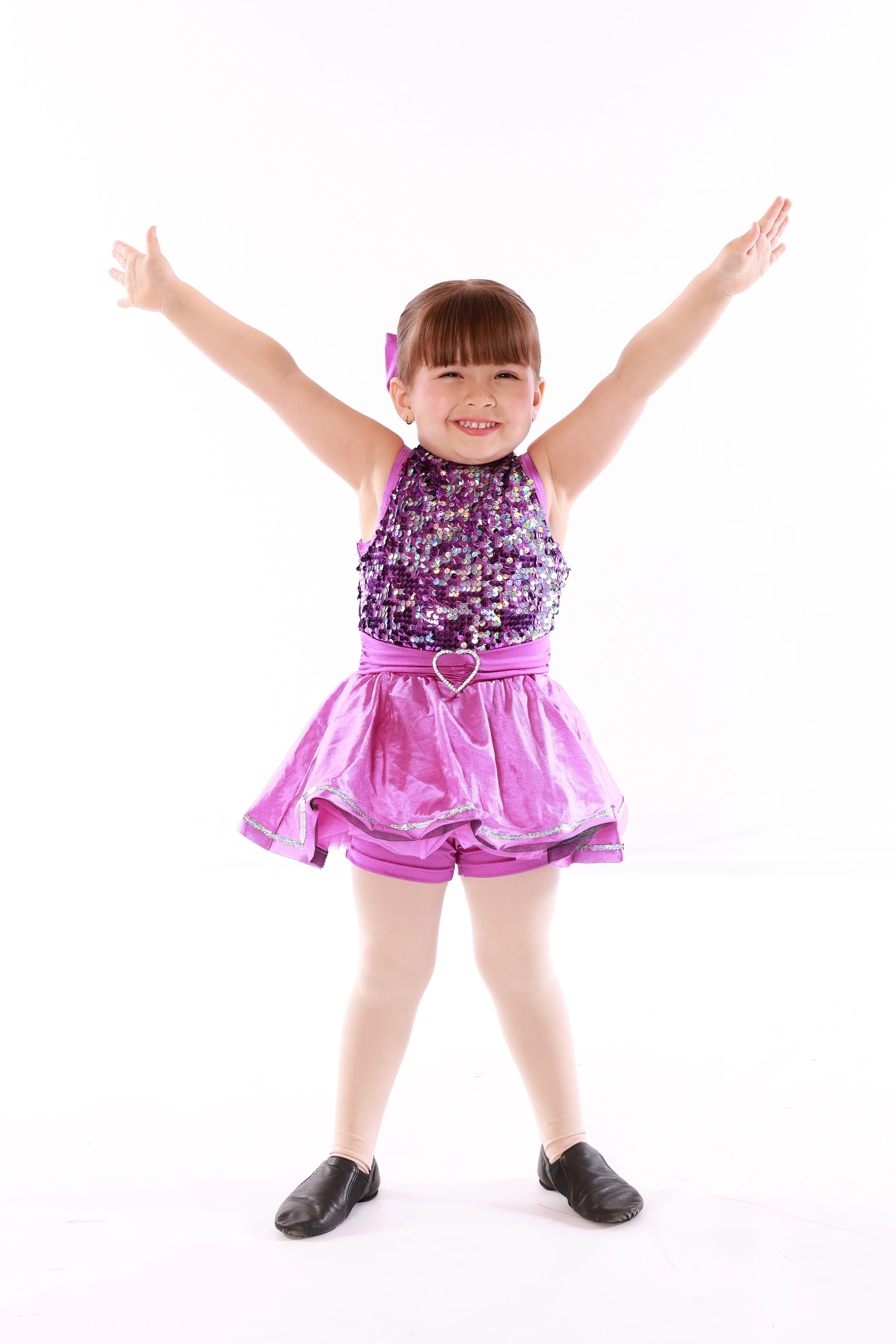 A young girl in a purple sequined dance costume with a pink tutu and tights, smiling with arms raised, standing on a white background.