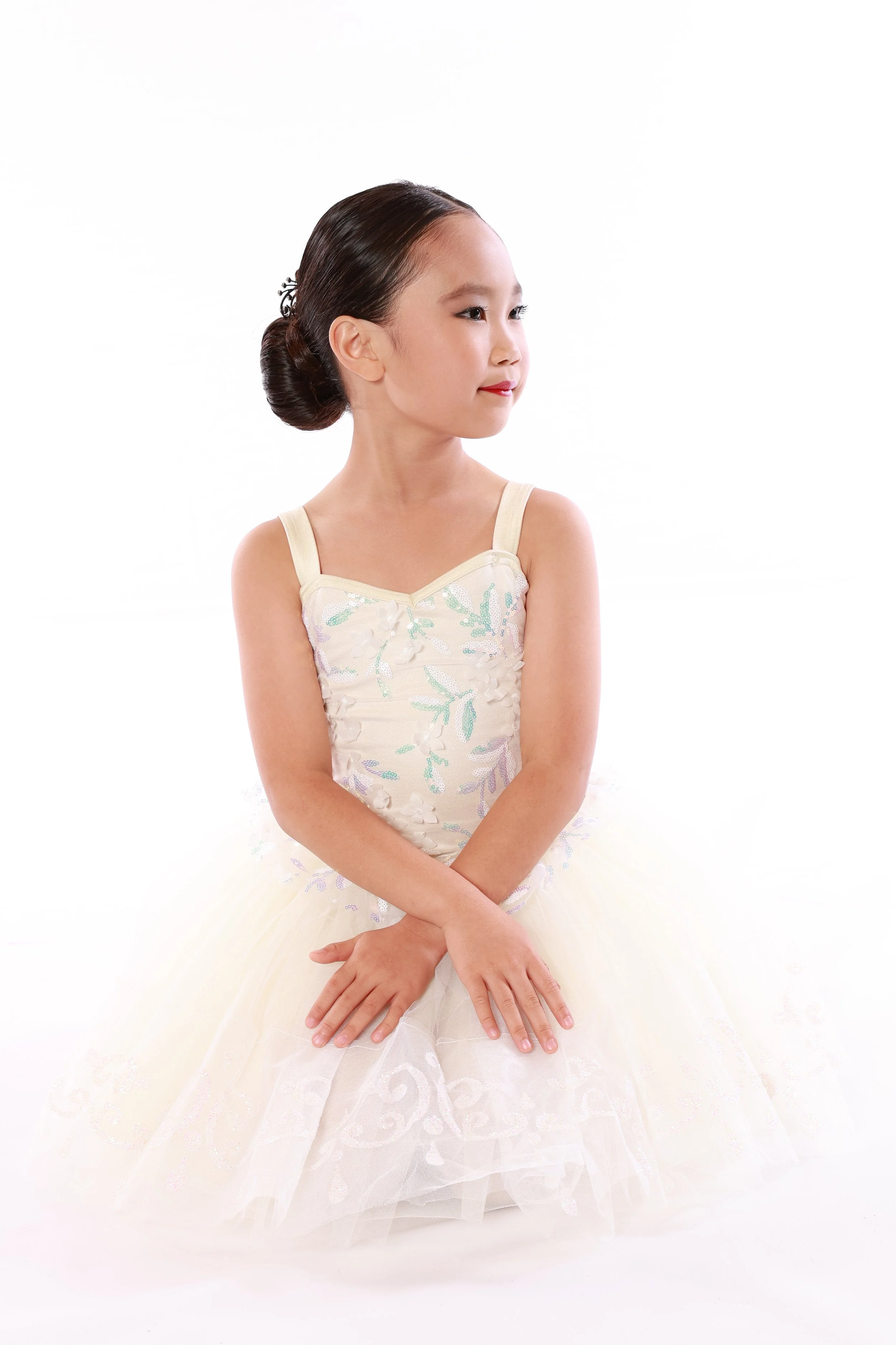 A young girl in a fancy dress, sitting with her hands folded on her lap, looking to the side, with a neutral expression.