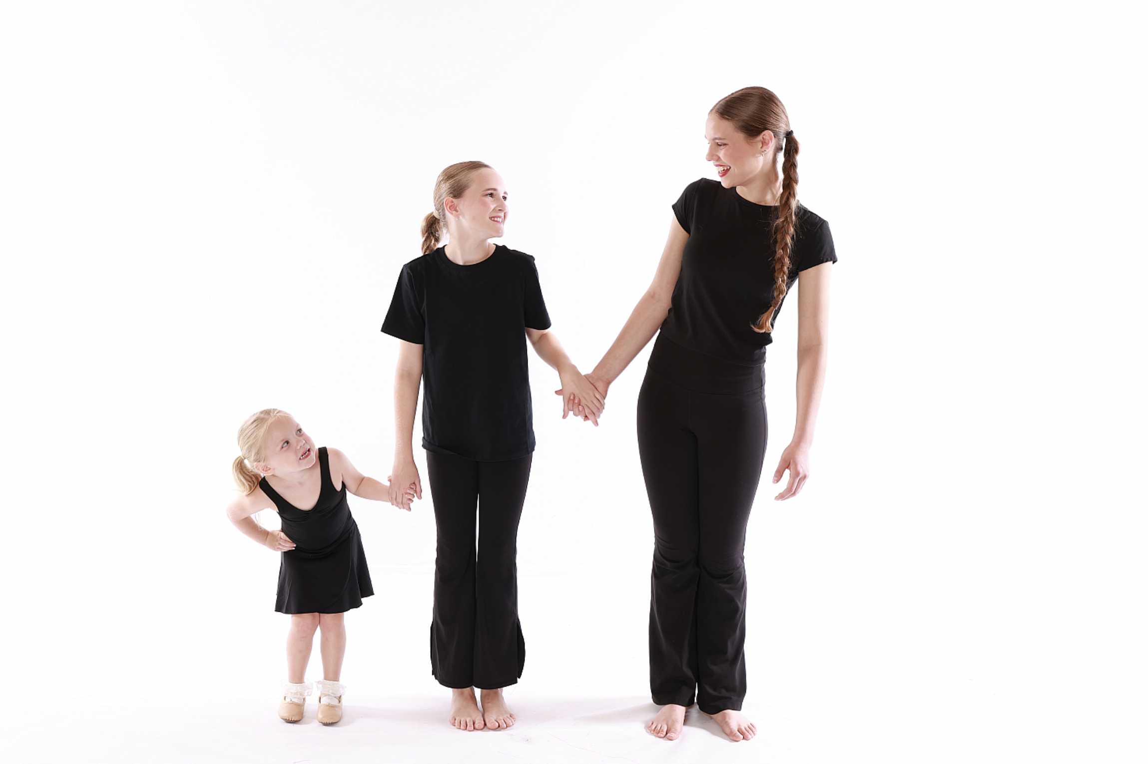 Three females, a toddler, a young girl, and an adult woman, holding hands and smiling at each other against a white background.
