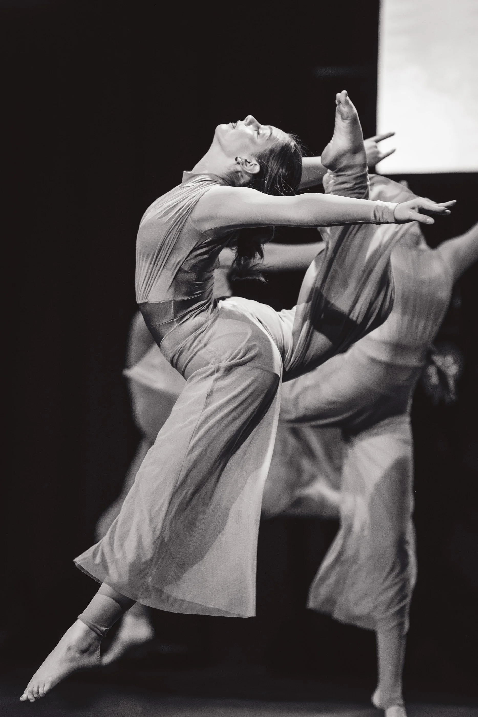 Ballet dancer performing a high arabesque with arms extended and head tilted back, in a flowy costume, black and white photograph.