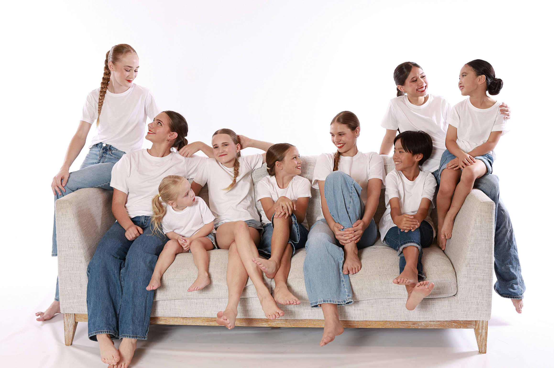 Ten girls of different ages sitting and standing on a beige sofa, wearing white t-shirts and jeans, smiling and talking with each other against a white background.