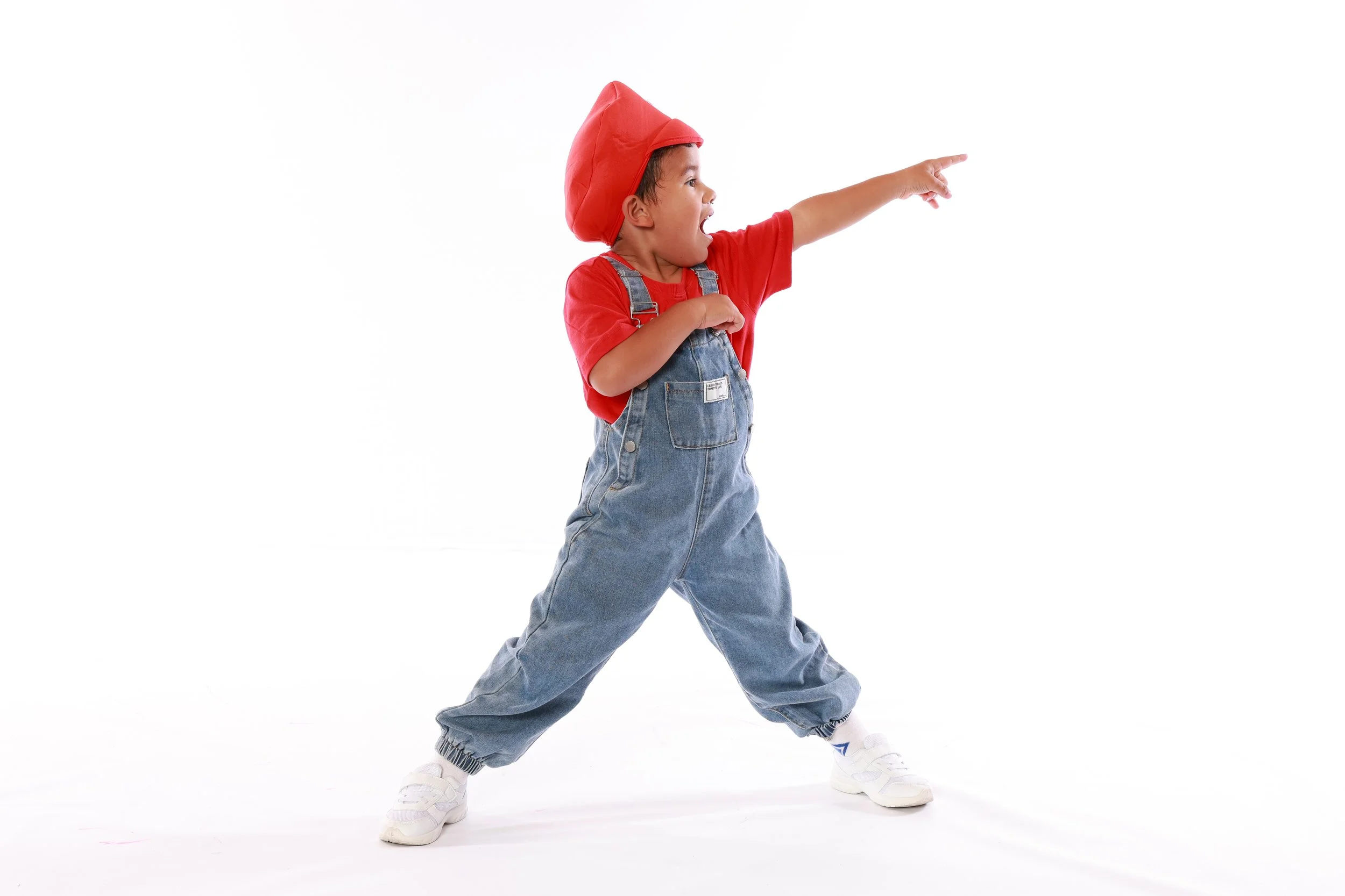 A young boy dressed in overalls, a red shirt, and a red hat, standing against a plain white background, pointing to the right with his mouth open.