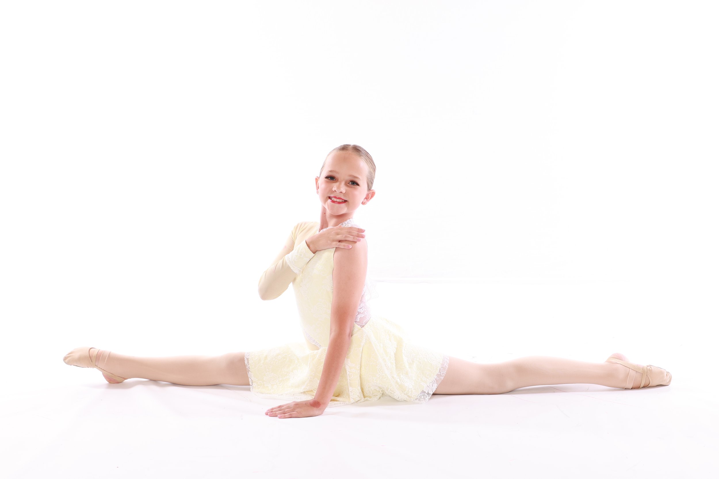 A young girl performing a split ballet pose on a white background, wearing a light yellow dance costume and ballet shoes, smiling at the camera.