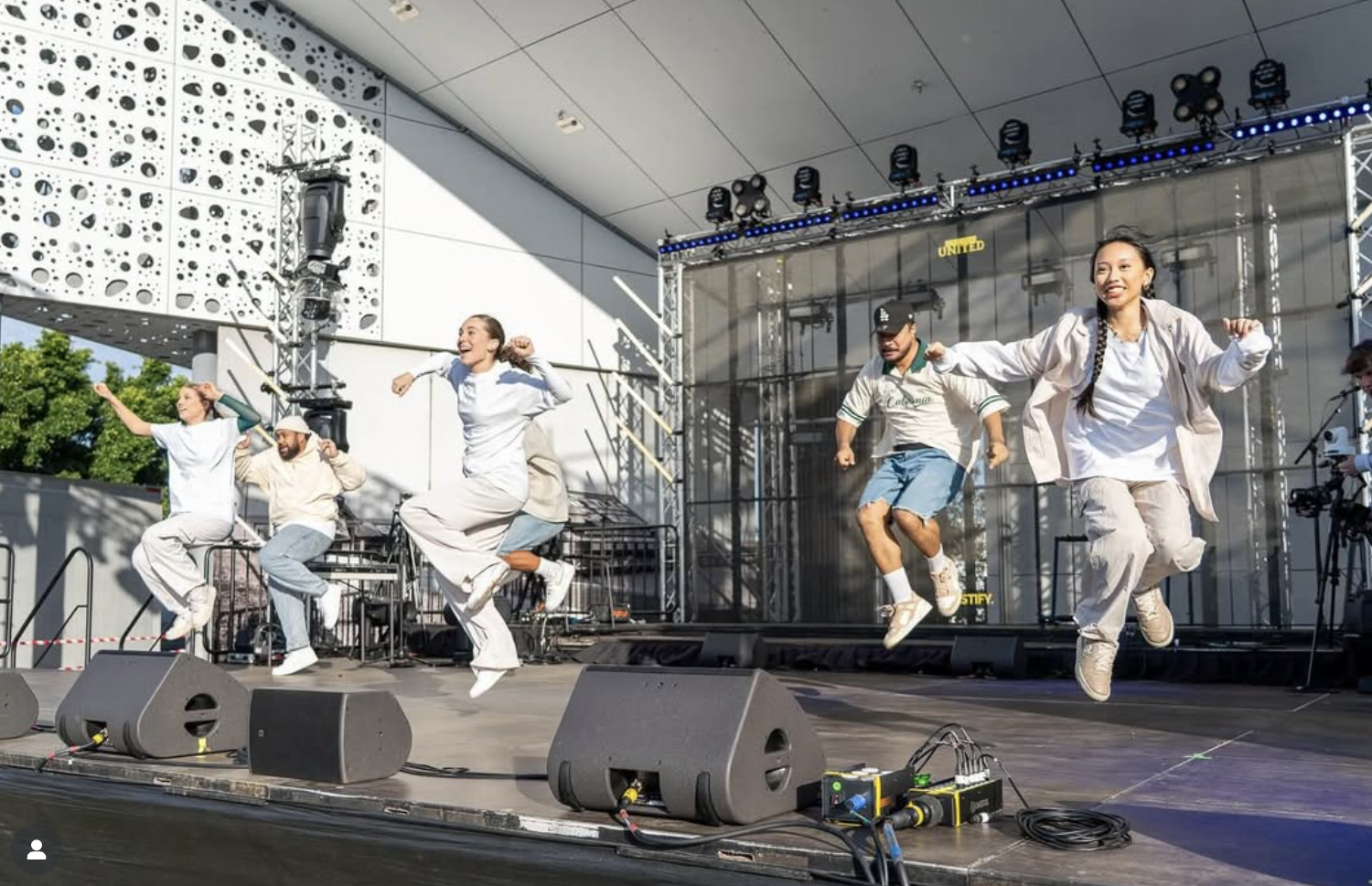 A group of six young people dancing and jumping on an outdoor stage with audio equipment and a modern building in the background during the daytime.