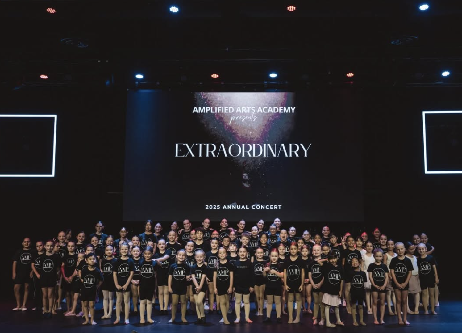 Group of young dancers on stage at Amplified Arts Academy's 2025 annual concert, with a large screen behind them displaying the event's theme 'Extraordinary'.