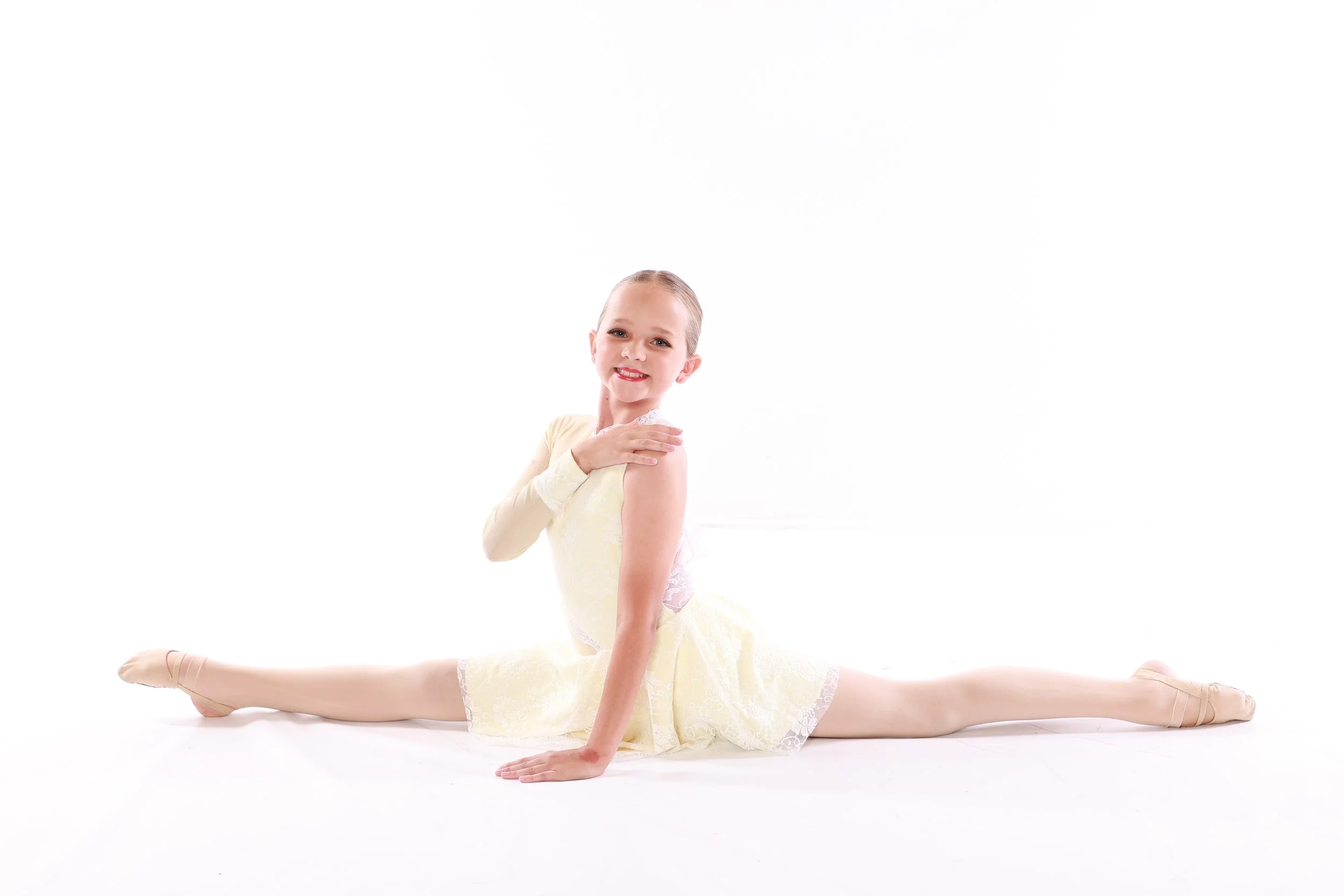 Young girl in a yellow ballet dress and tights performing a split dance move on a white background.
