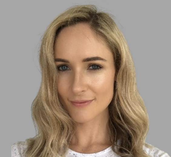 Portrait of a young woman with blonde wavy hair and blue eyes, smiling slightly, against a neutral gray background.
