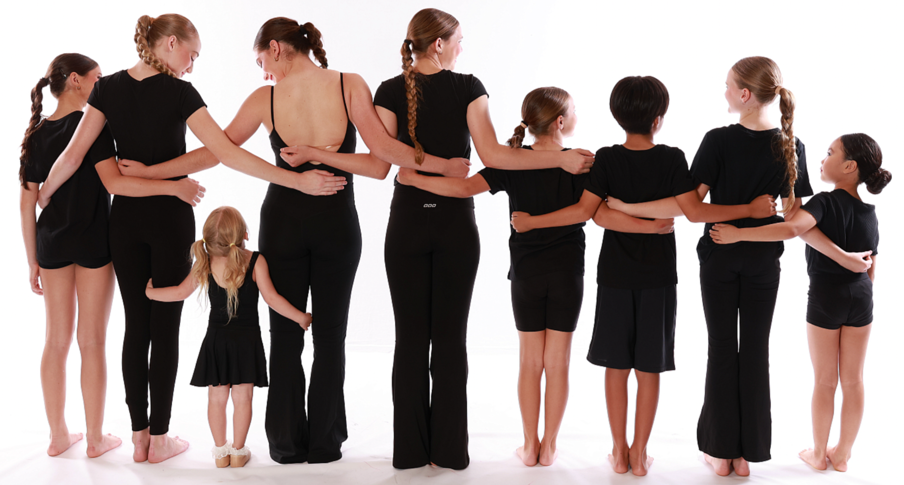 Group of nine females and one young girl standing together with their arms around each other, facing away, wearing black clothing against a white background.