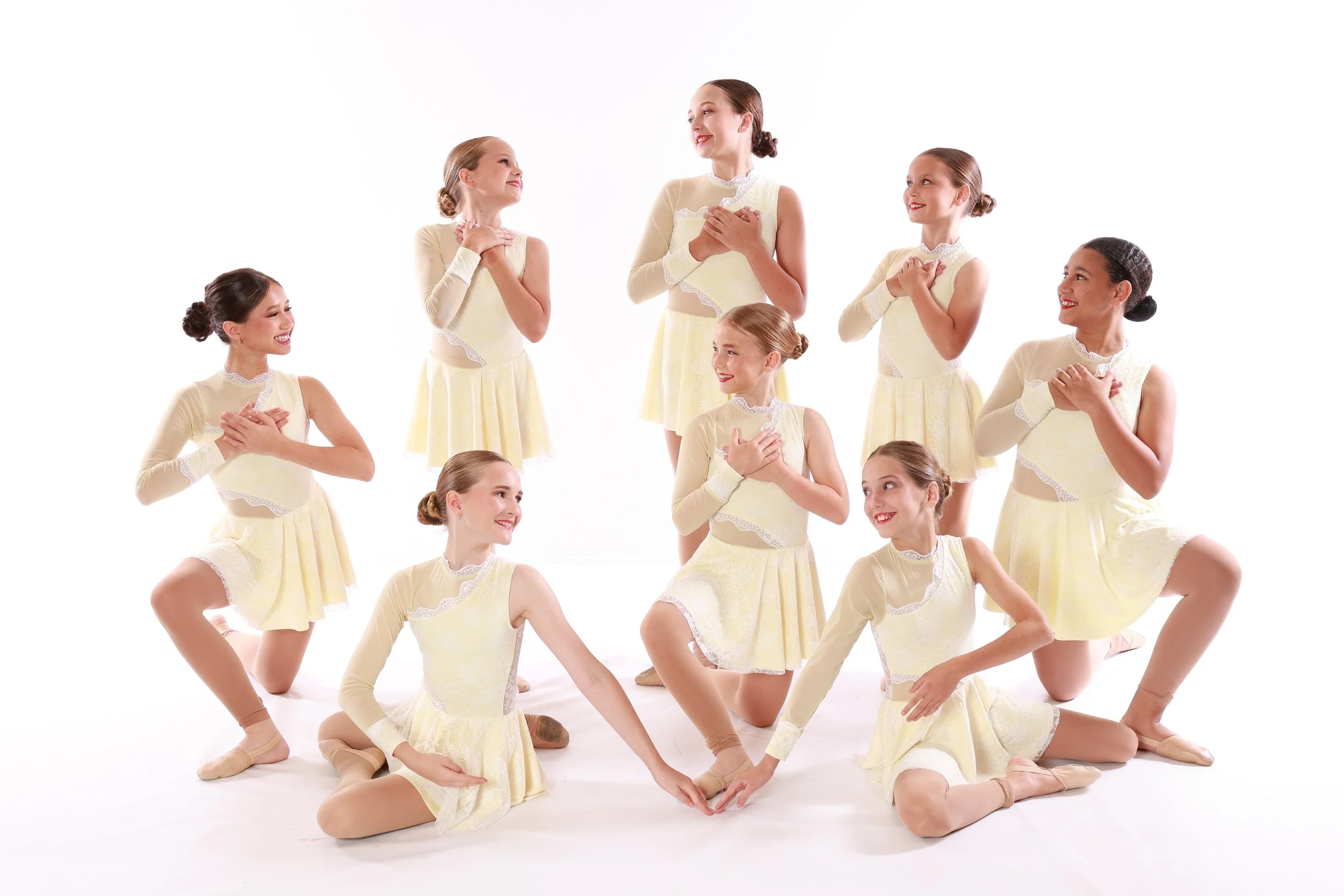 Group of young ballet dancers in yellow dresses and tights, smiling and looking at each other, in a ballet studio with a white background.