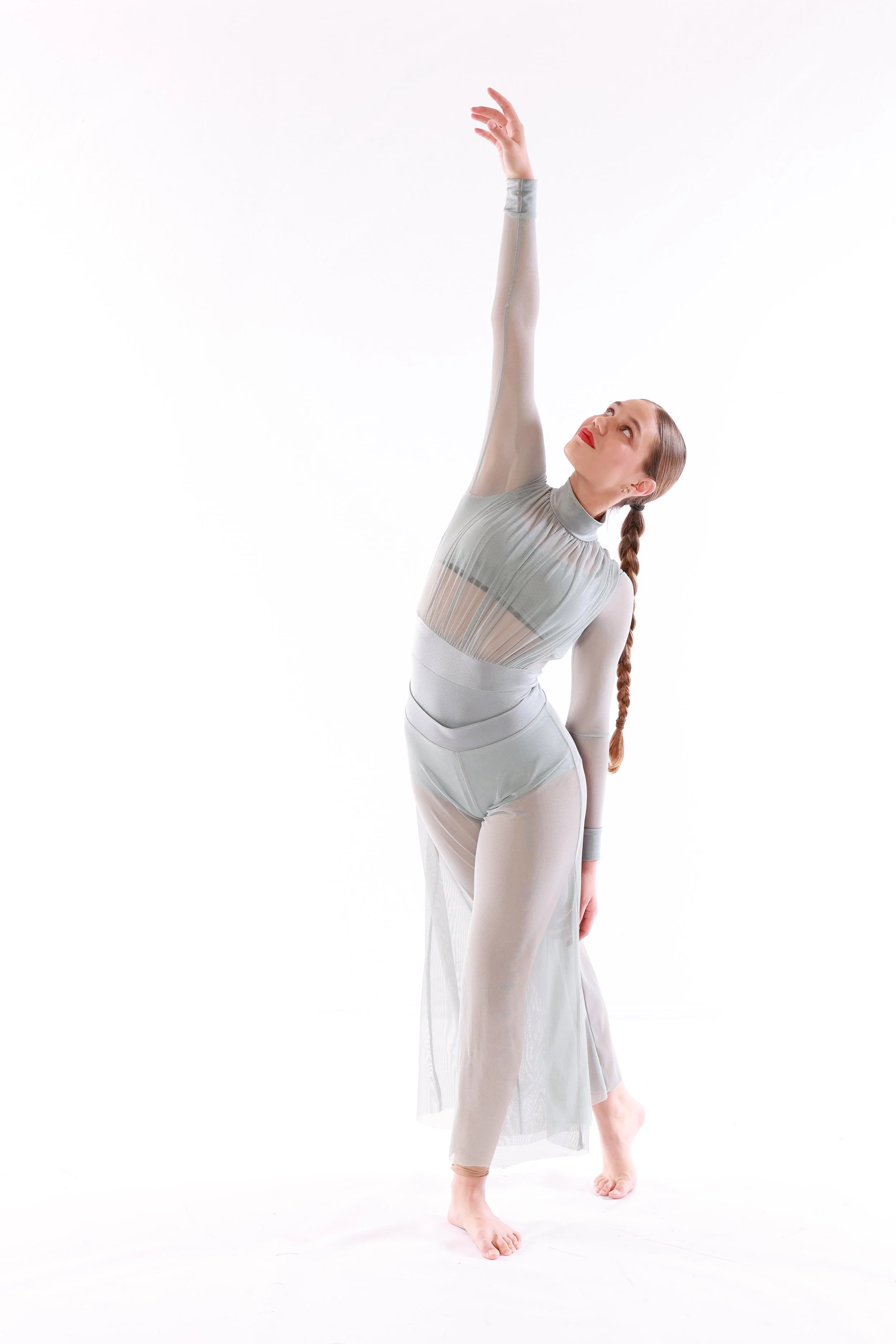 A woman in a sheer, light gray dance costume with a high neck, long sleeves, and wide-legged pants, is standing on one foot with her other leg bent and her toe pointed, raising one arm above her head and looking upwards.