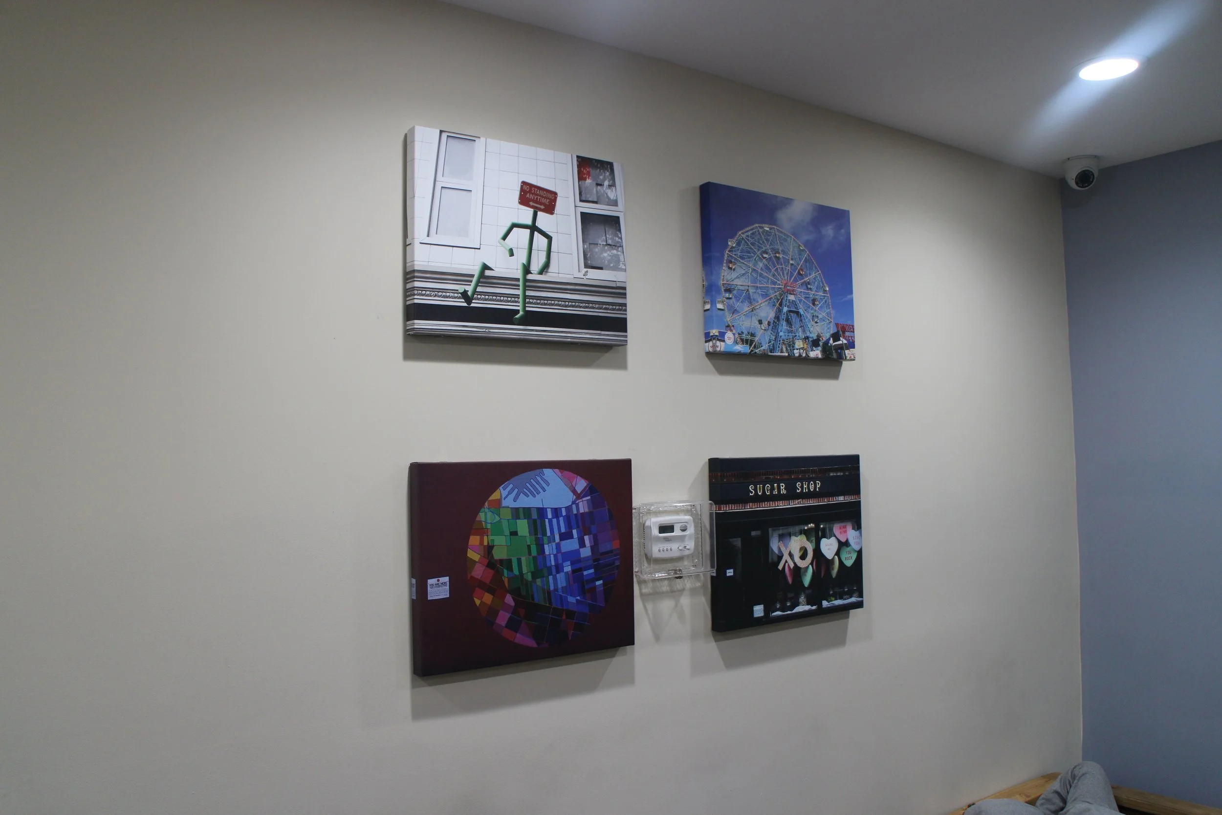 Five square pieces of wall art arranged in two rows, with three on the top row and two on the bottom, featuring a cityscape, Ferris wheel, abstract color pattern, and a candy shop sign, mounted on a beige wall in a room with ceiling lights and a secu