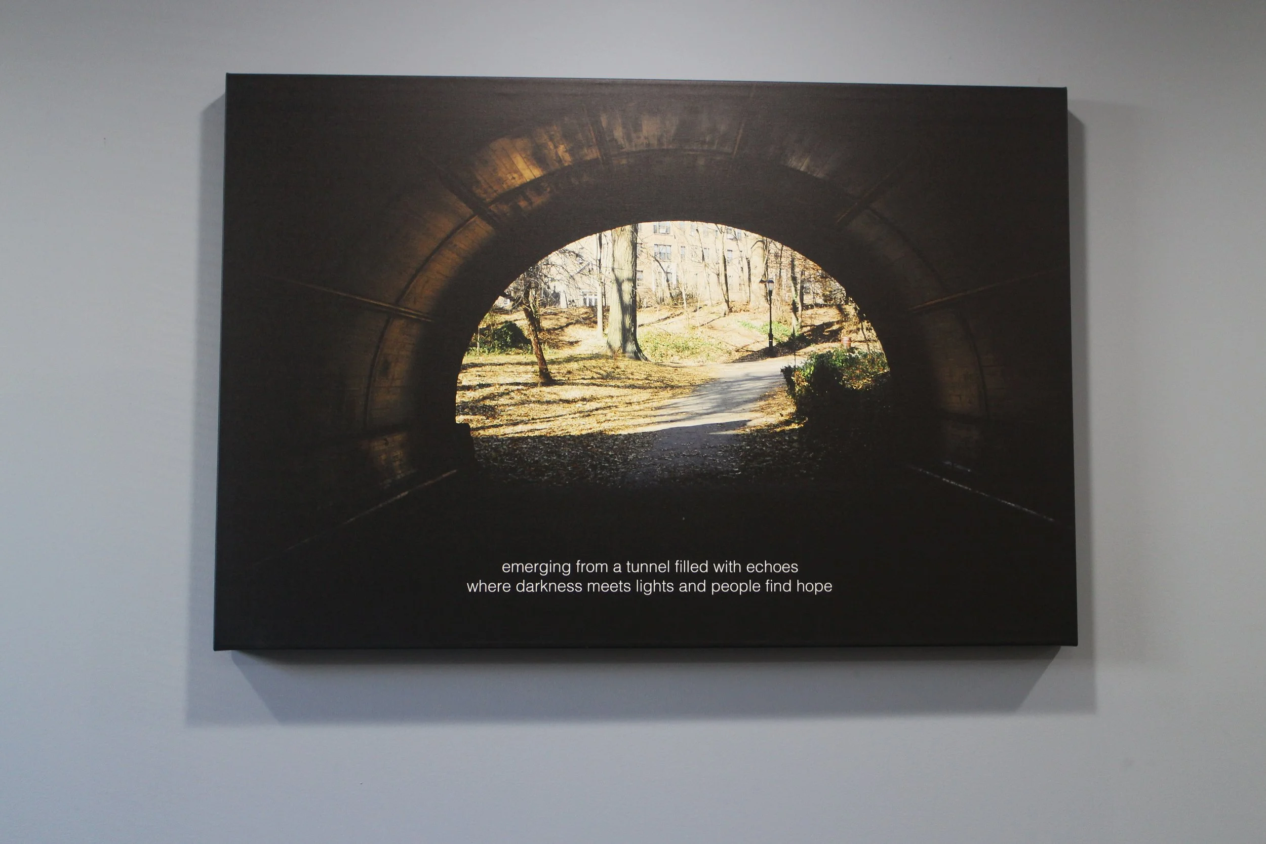 A photo of a tunnel with a view of a park and path outside, with sunlight illuminating trees and leaves, and a poem text at the bottom that reads: "emerging from a tunnel filled with echoes where darkness meets lights and people find hope."