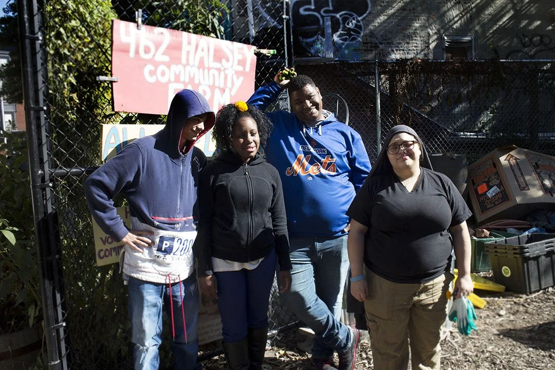 Four people standing outside in front of a fence with signs, including a young man with a Brooklyn Mets hoodie, a woman with glasses and black shirt, a young girl with curly hair and a black jacket, and a teen boy with a blue hoodie. It appears to be