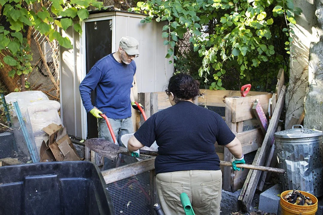 Two people working together on a backyard construction project, with one person shoveling dirt and the other holding a wheelbarrow.