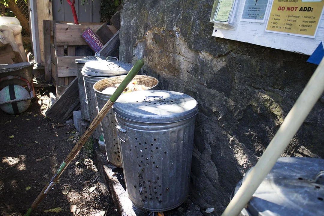 Two metal trash cans with lids, and a shovel with a green handle leaning against them, situated next to a stone wall and a wooden fence, in an outdoor setting.