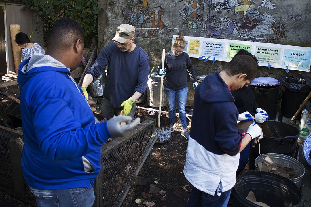 People participating in a community cleanup, sorting scrap metal and waste into containers, with a mural wall and cleanup instructions in the background.