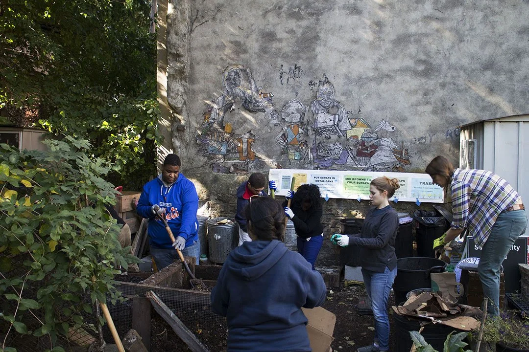 A group of people working together in a community cleanup effort, planting or tidying up a garden area next to a wall with a mural. The scene shows several volunteers, some with gloves and tools, cleaning and preparing the space.
