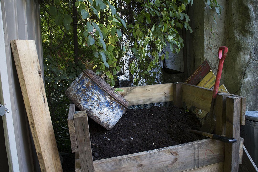 A wooden raised garden bed filled with dark soil, with gardening tools like a small shovel and a pruning saw, and a rusty metal bucket nearby, set outdoors with green leafy plants and a fence in the background.