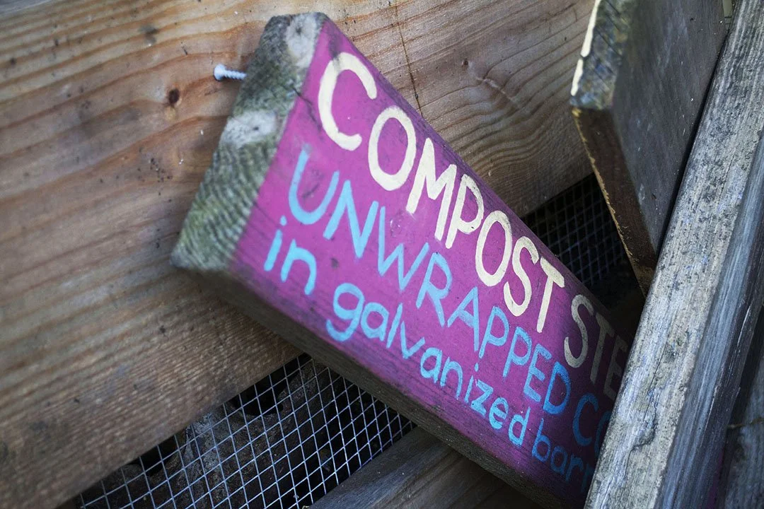 Handmade wooden sign painted pink and purple that reads, 'Compost Site Unwrapped Cart in galvanized bar' leaning against a wooden wall inside a fenced enclosure.