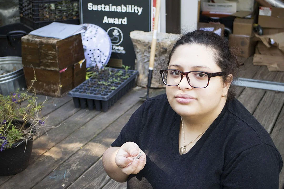 Young woman with glasses sitting on a wooden deck in front of a 'Sustainability Award' sign, holding a small plant seedling, surrounded by gardening supplies and plants.