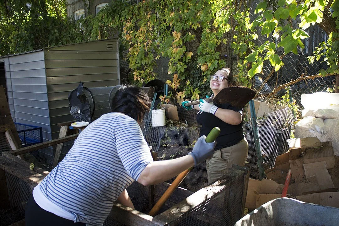Two women working together in a garden or compost area, one holding a gardening tool and the other holding a trowel, surrounded by plants, trees, and gardening supplies.